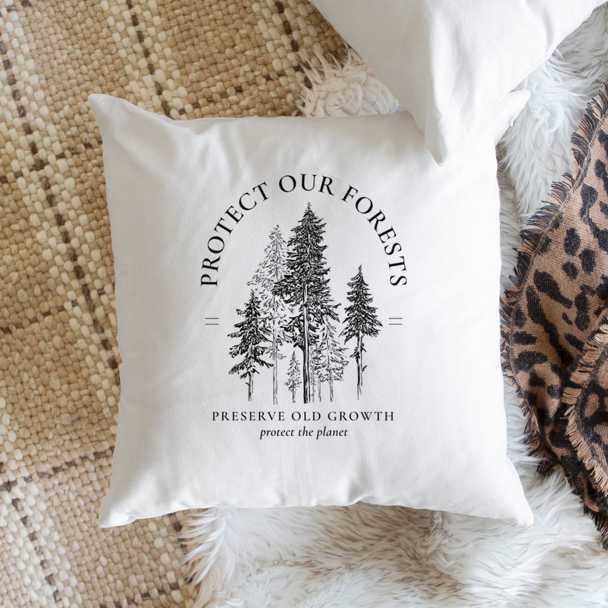 White pillow cover printed with a black ink illustration of a stand of tall pine trees, framed by the words Protect Our Forests, Preserve Old Growth, on a woven chair.