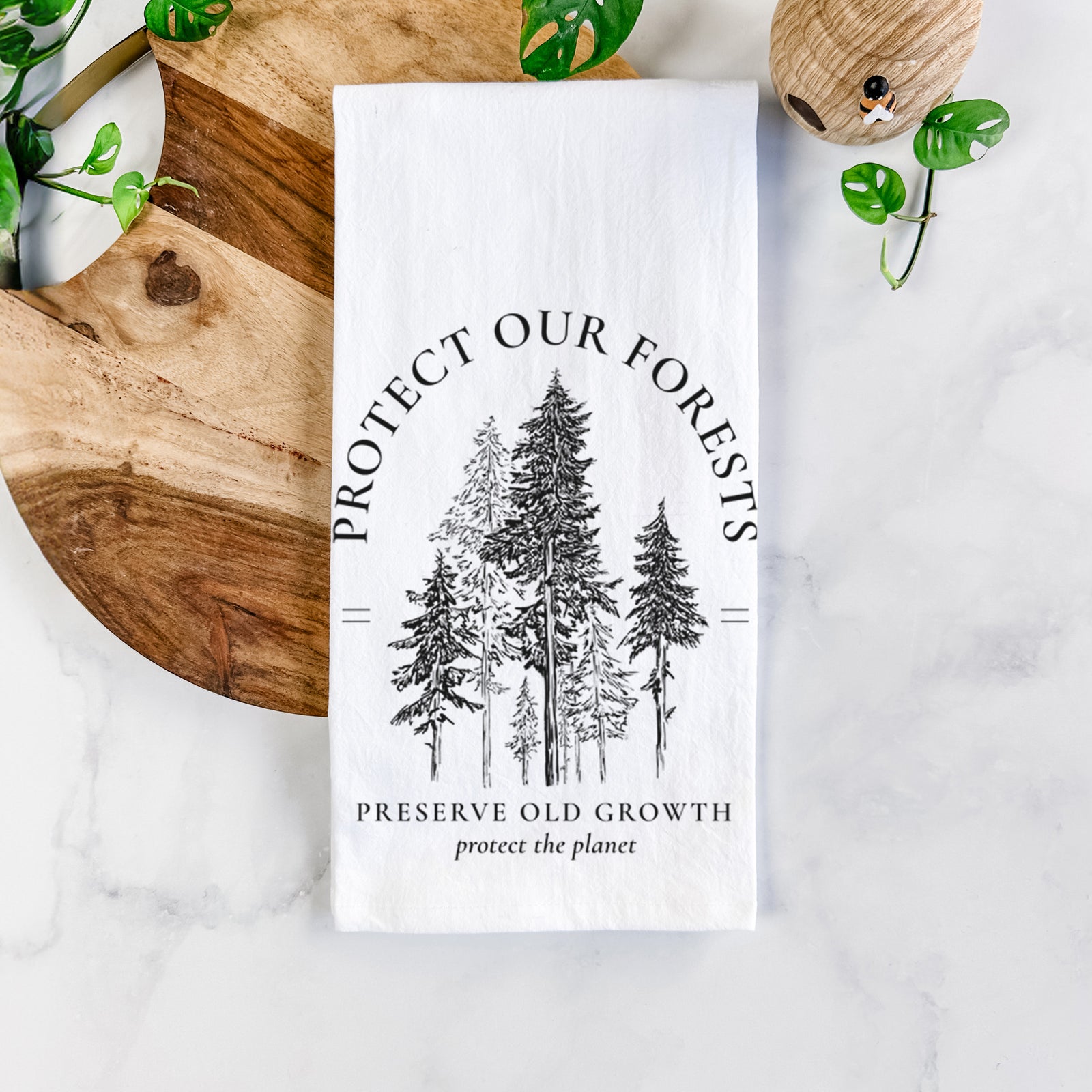 White cotton tea towel printed with a black ink illustration of a stand of tall pine trees, framed by the words Protect Our Forests, Preserve Old Growth, on a marble counter beside a round wooden cutting board and greenery.