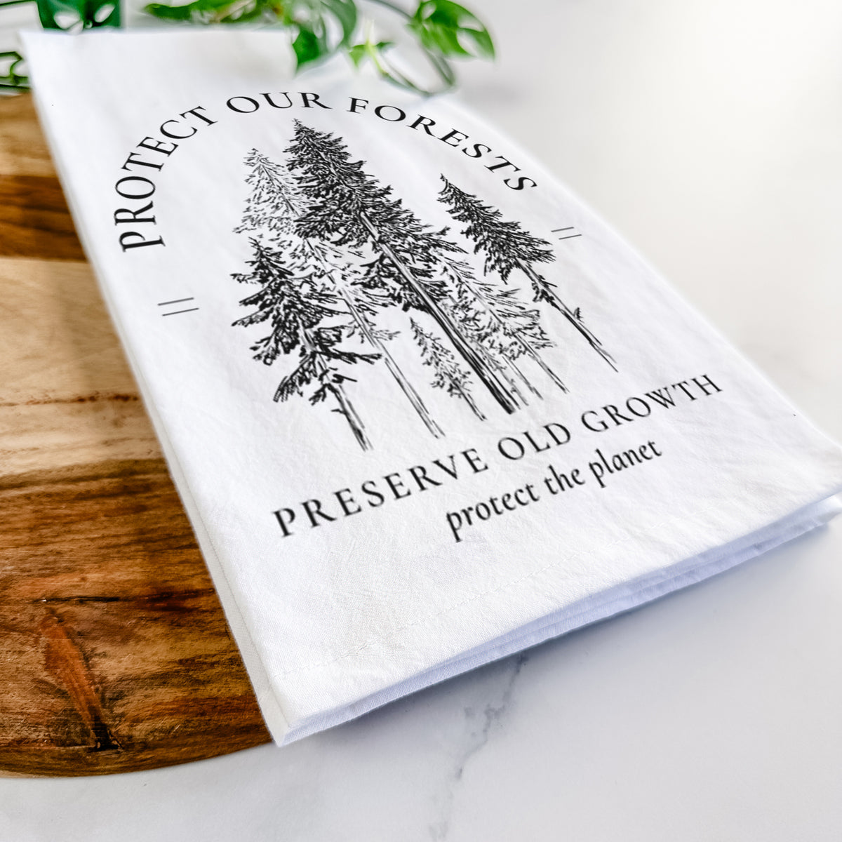 Close-up of a folded white tea towel on a wooden board, showing a black ink illustration of a stand of tall pine trees, framed by the words Protect Our Forests, Preserve Old Growth.