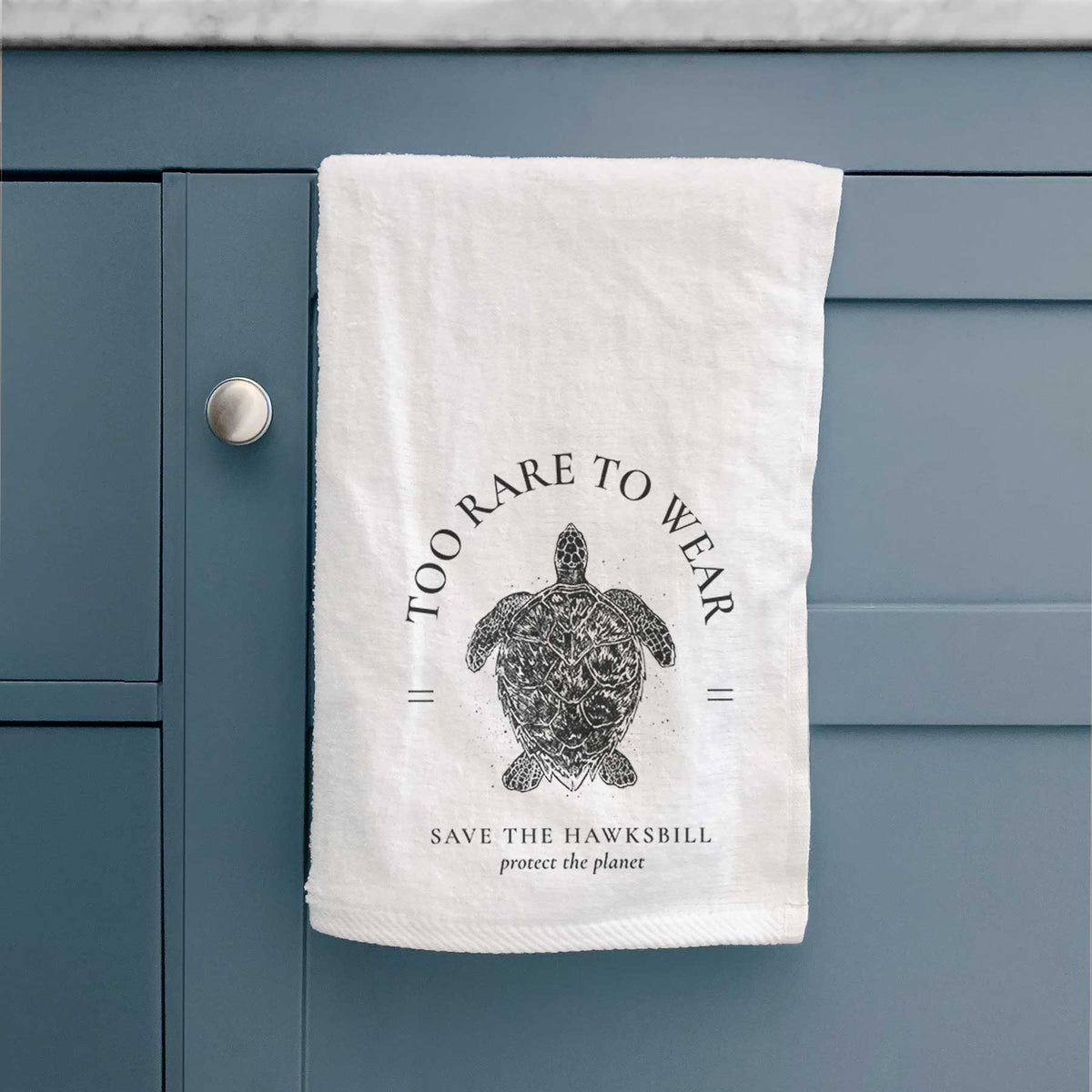 White hand towel printed with a black ink hawksbill sea turtle framed by the words Too Rare to Wear, Save the Hawksbill, hanging on a cabinet door.