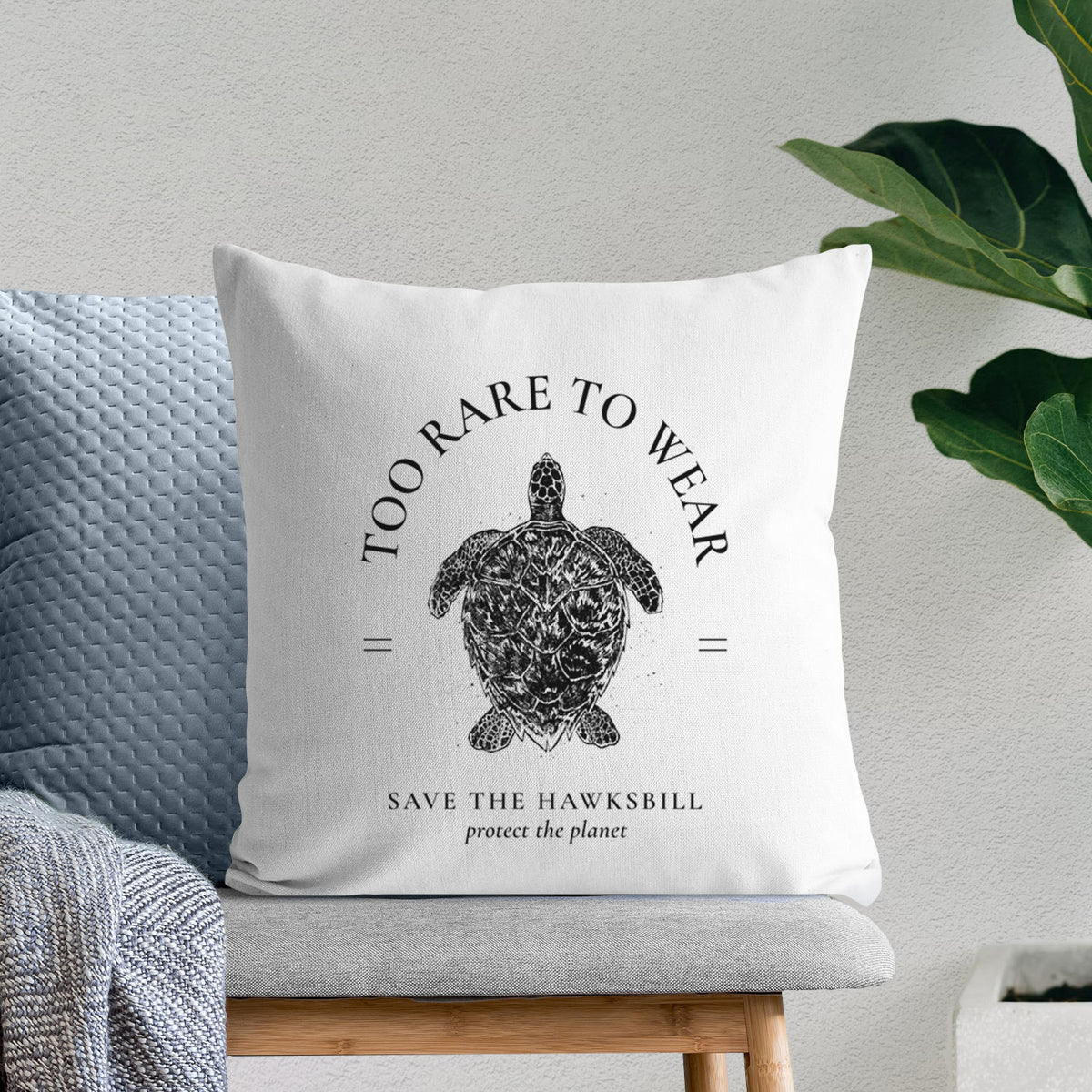 White pillow cover printed with a black ink hawksbill sea turtle framed by the words Too Rare to Wear, Save the Hawksbill, on a bench beside plants.