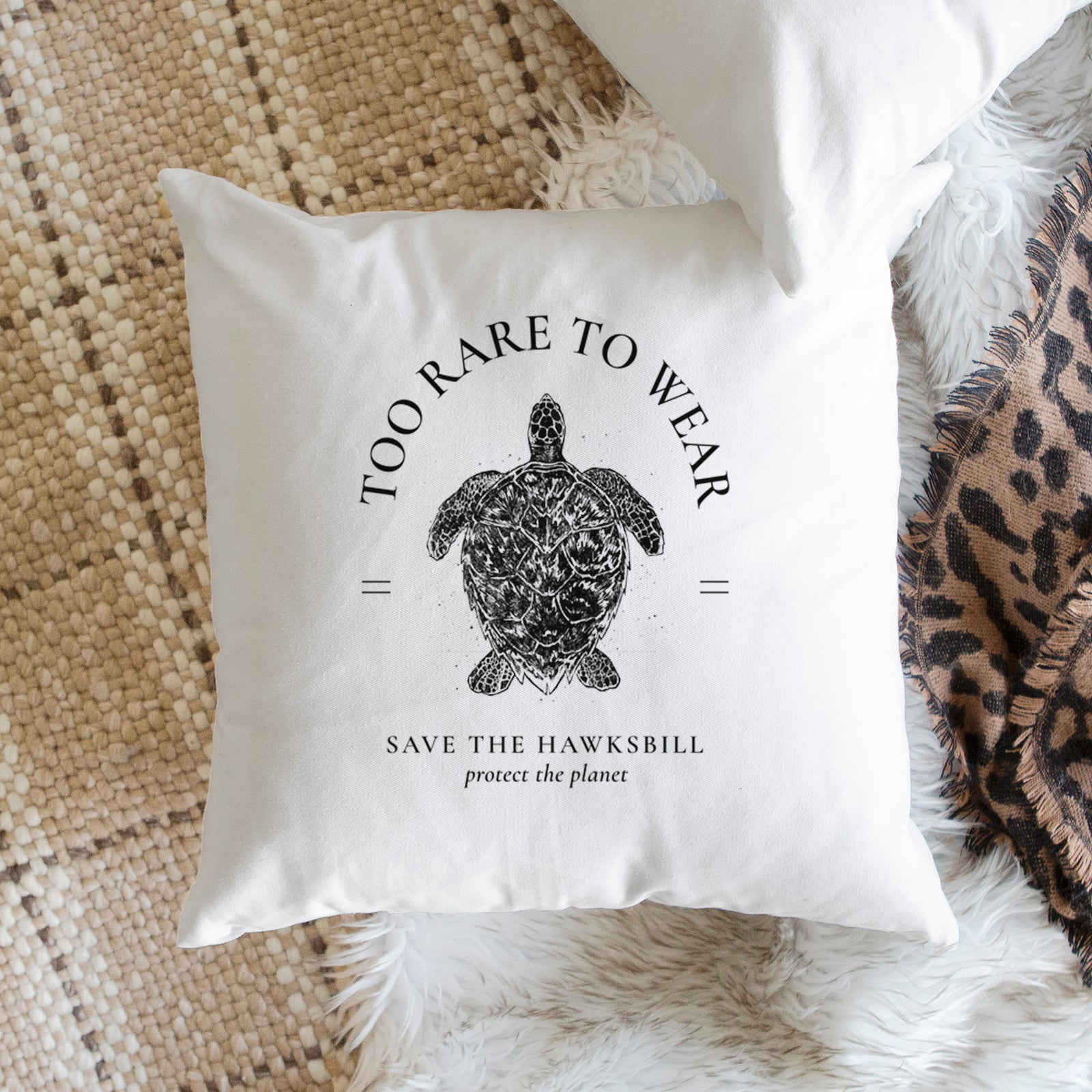 White pillow cover printed with a black ink hawksbill sea turtle framed by the words Too Rare to Wear, Save the Hawksbill, on a bench beside plants.