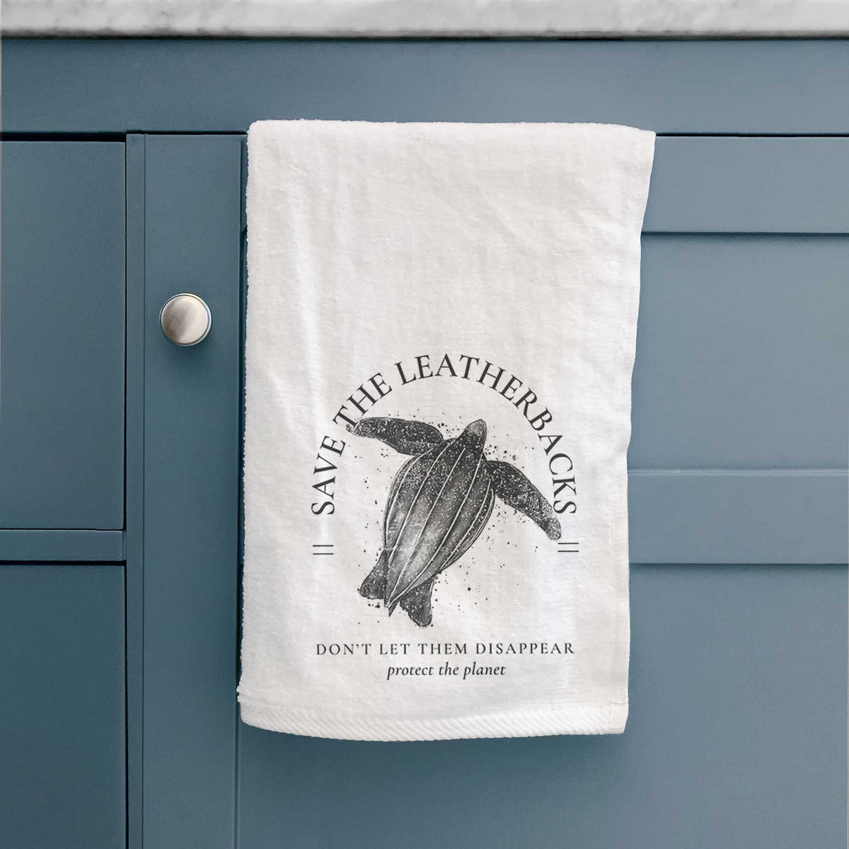 White hand towel printed with a black ink leatherback sea turtle framed by the words Save the Leatherbacks, Don't Let Them Disappear, hanging on a cabinet door.