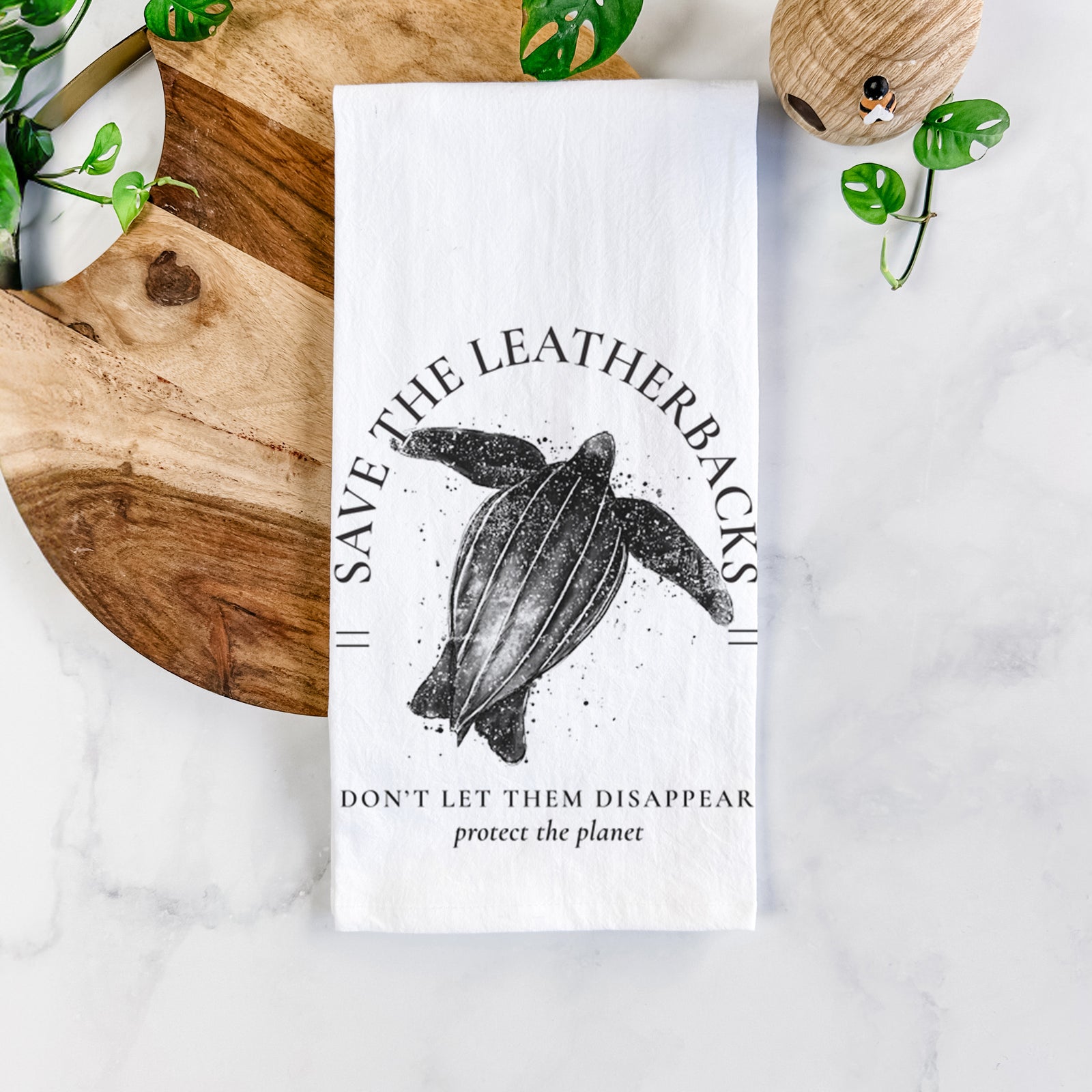White cotton tea towel printed with a black ink leatherback sea turtle framed by the words Save the Leatherbacks, Don't Let Them Disappear, on a marble counter beside a round wooden cutting board and greenery.
