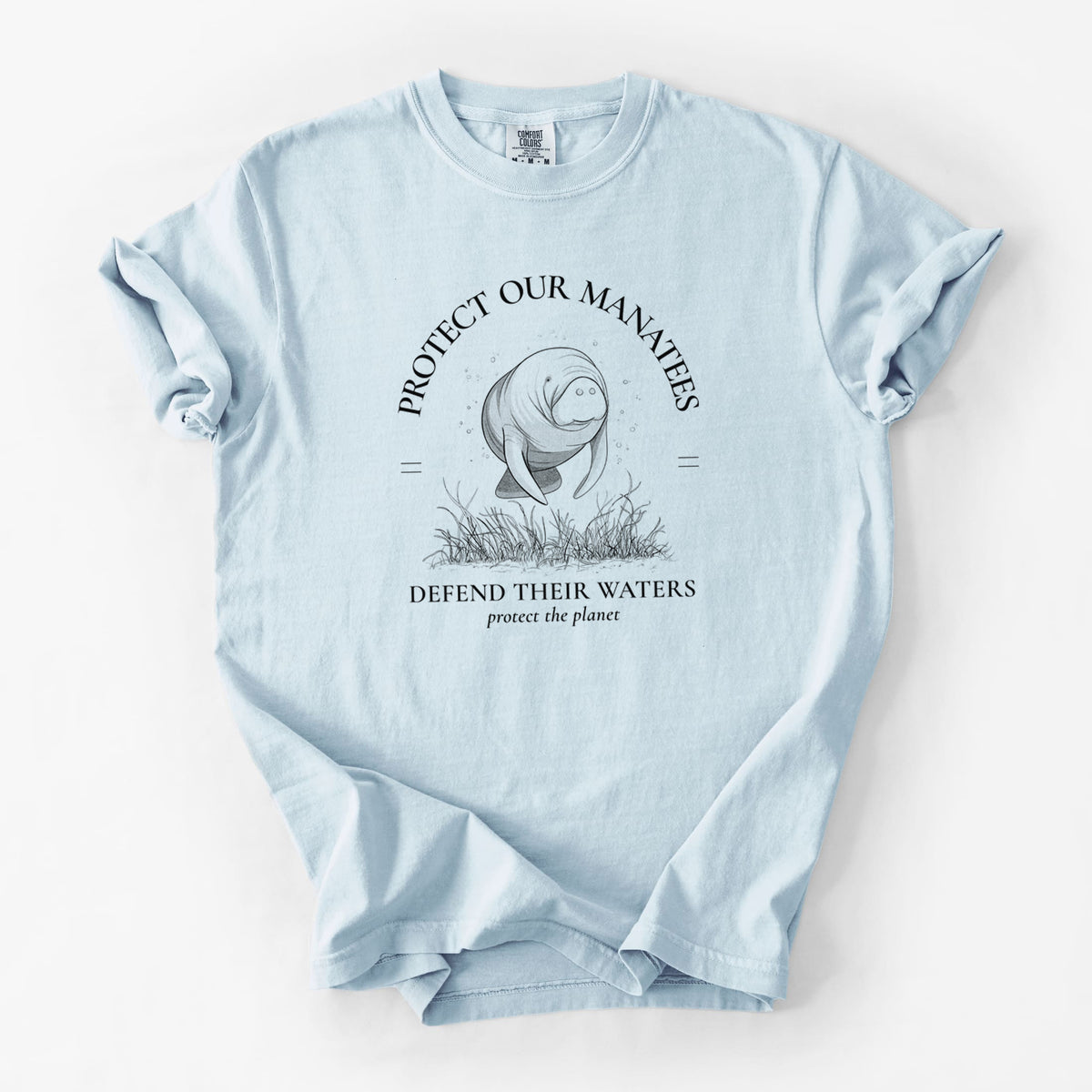 Chambray blue Comfort Colors t-shirt laid flat, printed with a black ink illustration of a manatee among seagrass, framed by the words Protect Our Manatees, Defend Their Waters, Protect the Planet.