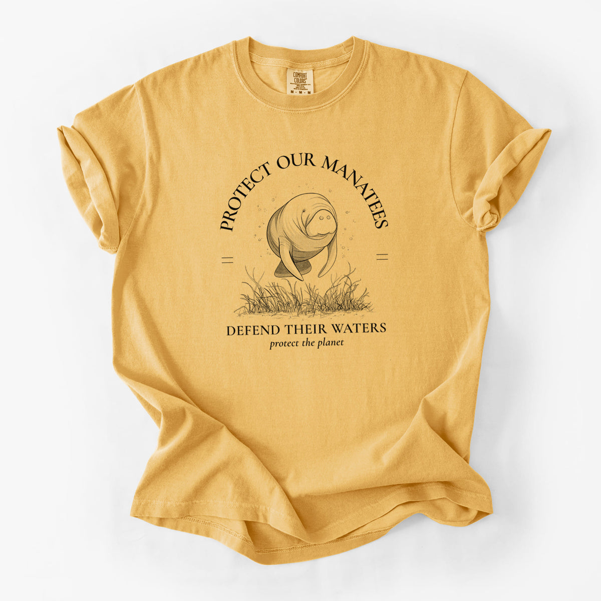 Golden wheat yellow Comfort Colors t-shirt laid flat, printed with a black ink illustration of a manatee among seagrass, framed by the words Protect Our Manatees, Defend Their Waters, Protect the Planet.