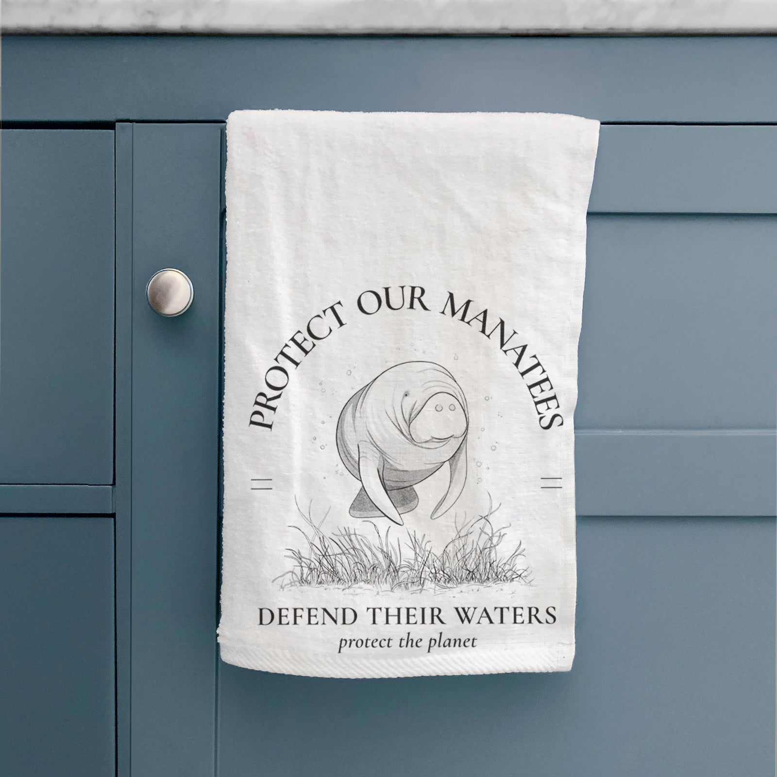 White hand towel printed with a black ink illustration of a manatee among seagrass, framed by the words Protect Our Manatees, Defend Their Waters, Protect the Planet.