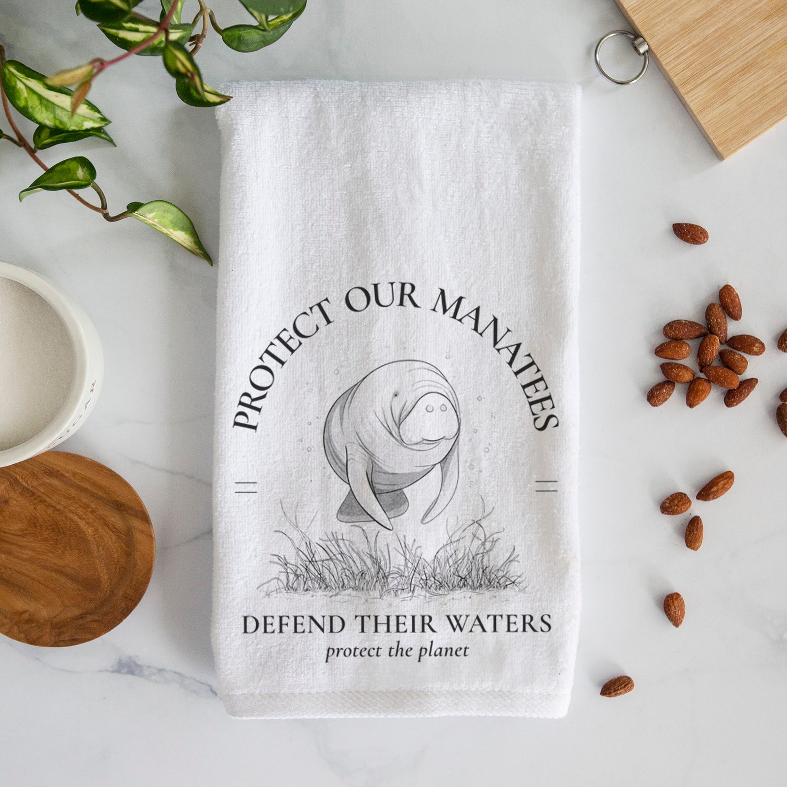 White hand towel printed with a black ink illustration of a manatee among seagrass, framed by the words Protect Our Manatees, Defend Their Waters, Protect the Planet.