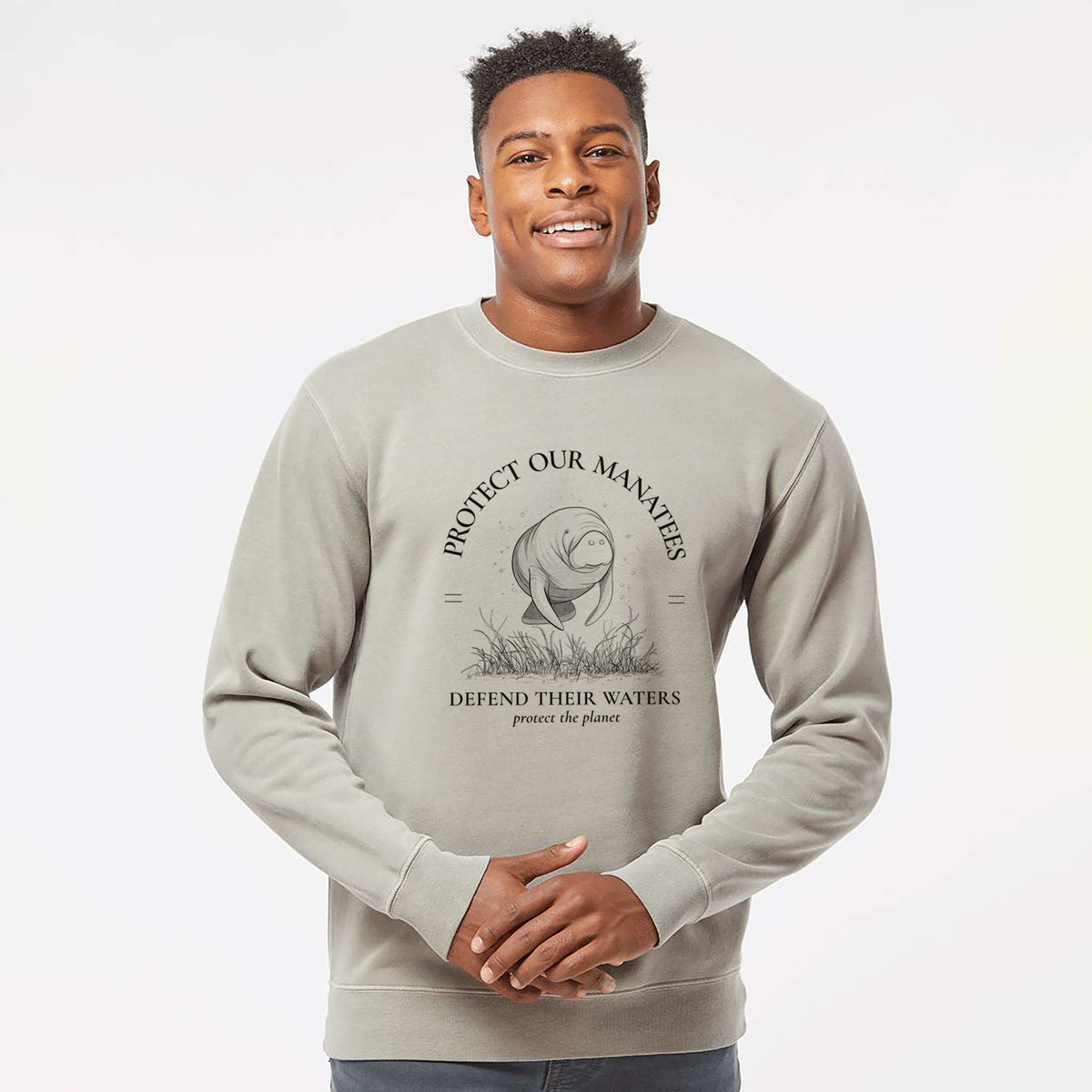Model wearing a cement grey pigment dyed crewneck sweatshirt printed with a black ink illustration of a manatee among seagrass, framed by the words Protect Our Manatees, Defend Their Waters, Protect the Planet.