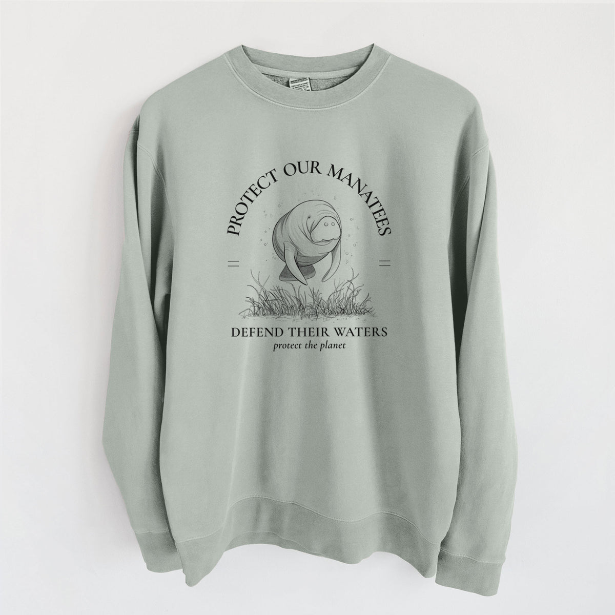 Sage green pigment dyed crewneck sweatshirt laid flat, printed with a black ink illustration of a manatee among seagrass, framed by the words Protect Our Manatees, Defend Their Waters, Protect the Planet.