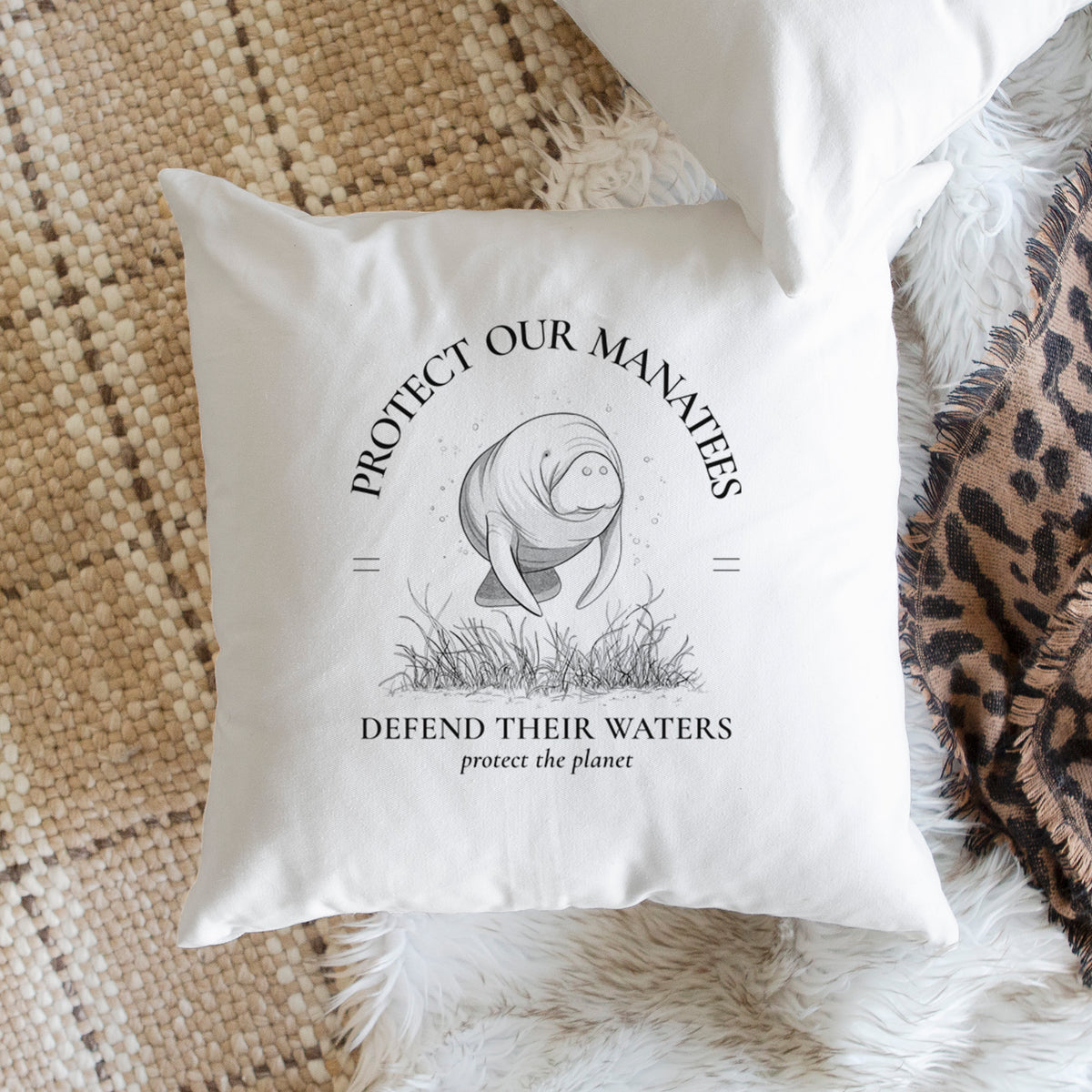 White pillow cover printed with a black ink illustration of a manatee among seagrass, framed by the words Protect Our Manatees, Defend Their Waters, Protect the Planet, on a woven chair.