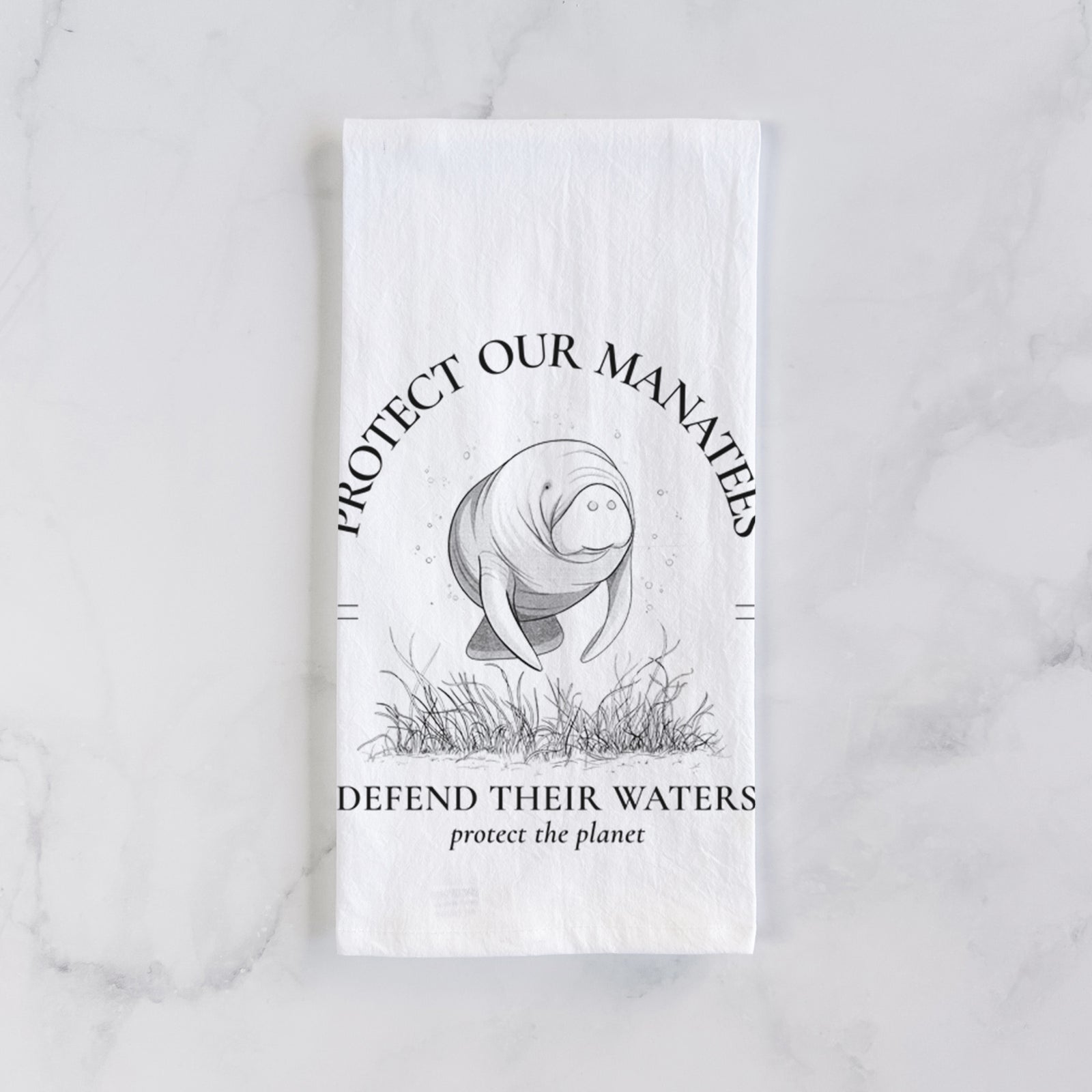 White cotton tea towel printed with a black ink illustration of a manatee among seagrass, framed by the words Protect Our Manatees, Defend Their Waters, Protect the Planet, on a marble counter beside a round wooden cutting board and greenery.