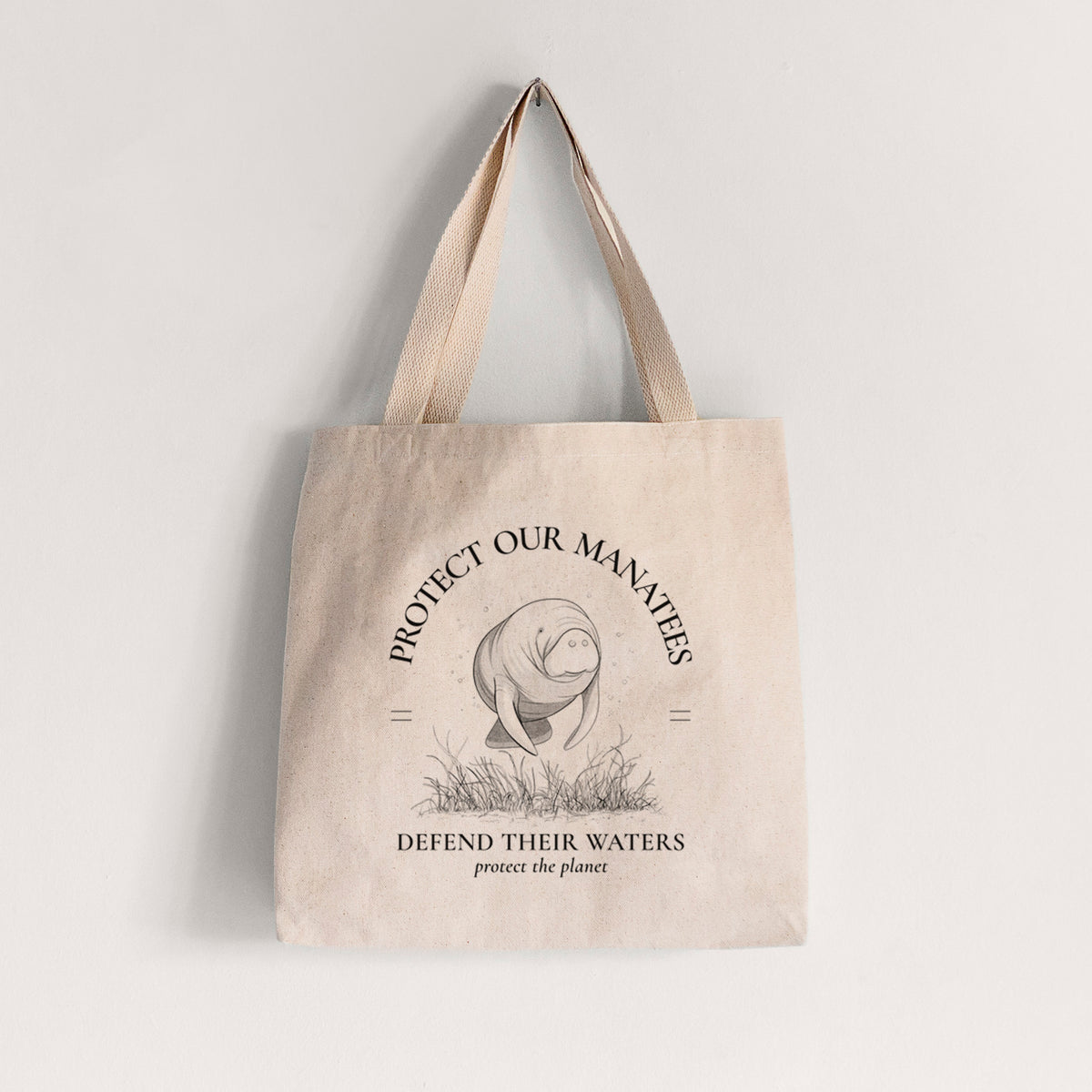 Natural canvas tote bag printed with a black ink illustration of a manatee among seagrass, framed by the words Protect Our Manatees, Defend Their Waters, Protect the Planet.