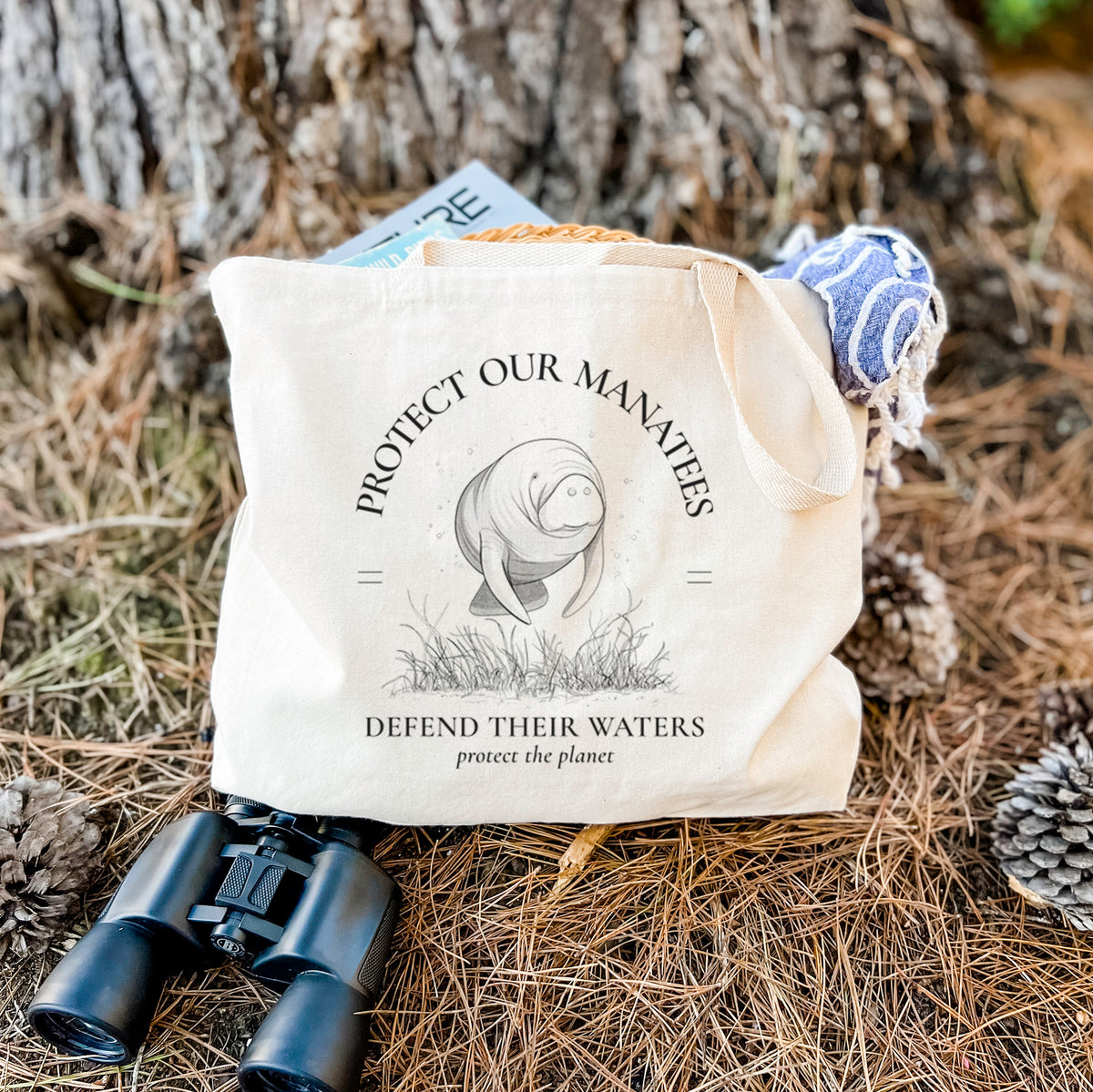 Canvas tote bag printed with a black ink illustration of a manatee among seagrass, framed by the words Protect Our Manatees, Defend Their Waters, Protect the Planet, resting on dry grass beside a pair of binoculars.