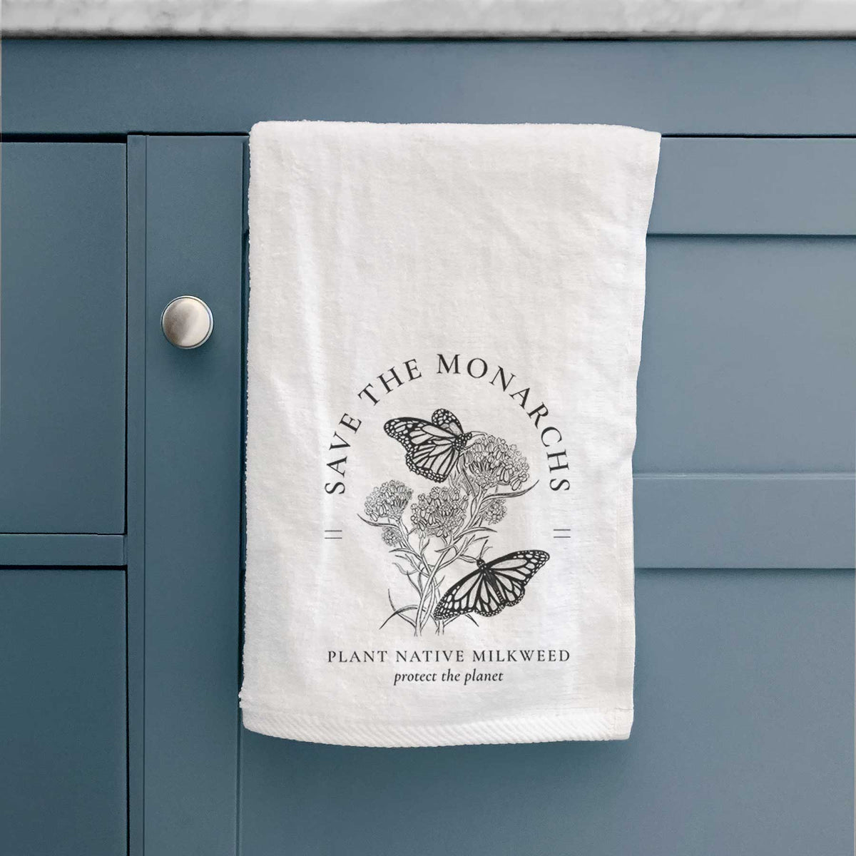 White hand towel printed with black ink monarch butterflies on milkweed flowers, framed by the words Save the Monarchs, Plant Native Milkweed, hanging on a cabinet door.