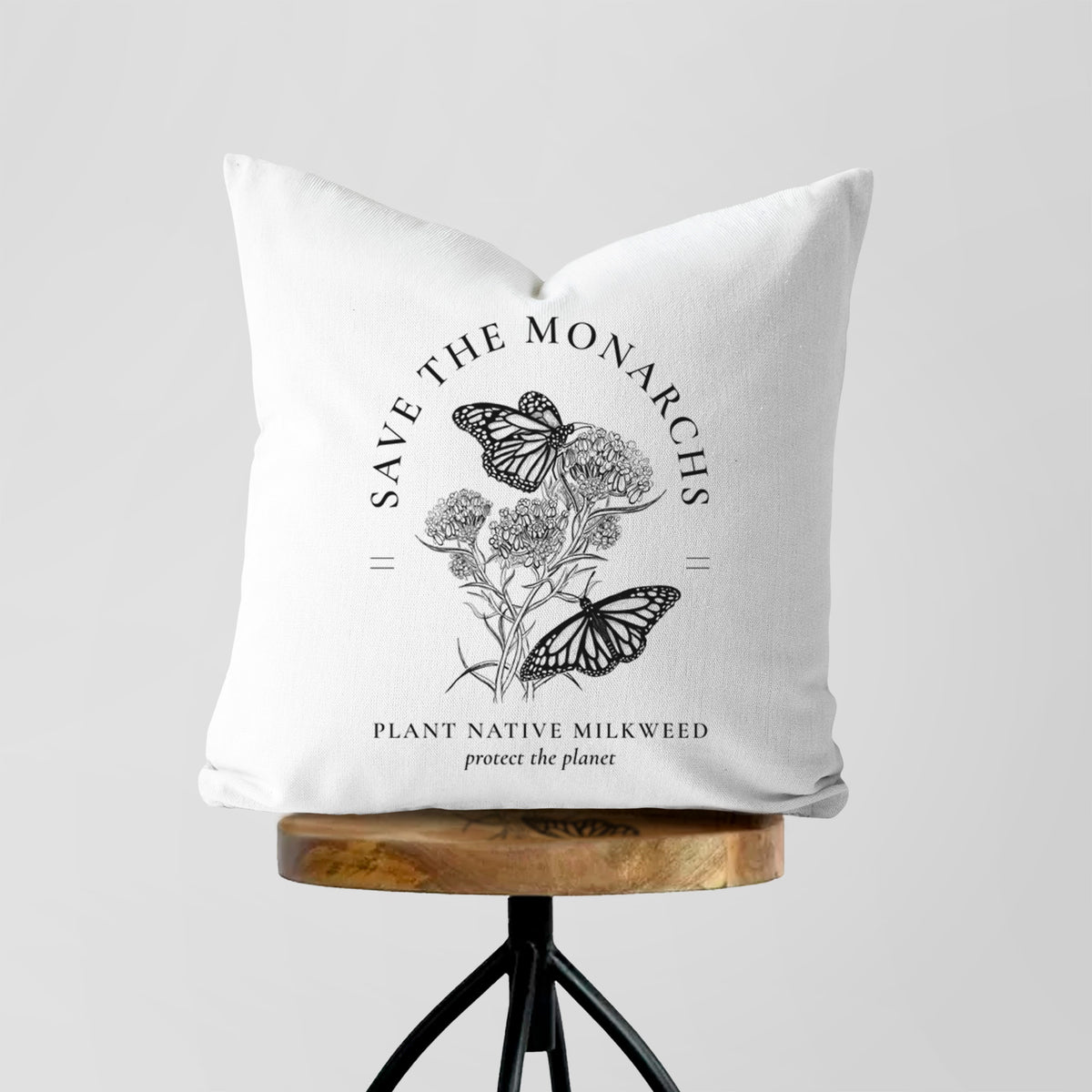 White pillow cover printed with black ink monarch butterflies on milkweed flowers, framed by the words Save the Monarchs, Plant Native Milkweed, on a wooden stool.