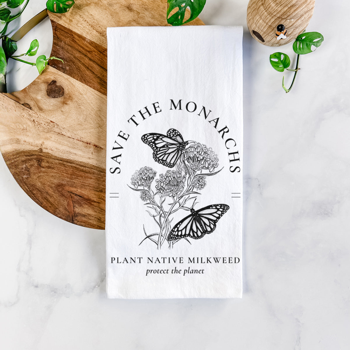 White cotton tea towel printed with black ink monarch butterflies on milkweed flowers, framed by the words Save the Monarchs, Plant Native Milkweed, on a marble counter beside a round wooden cutting board and greenery.
