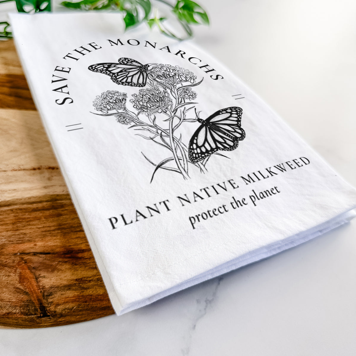 Close-up of a folded white tea towel on a wooden board, showing black ink monarch butterflies on milkweed flowers, framed by the words Save the Monarchs, Plant Native Milkweed.