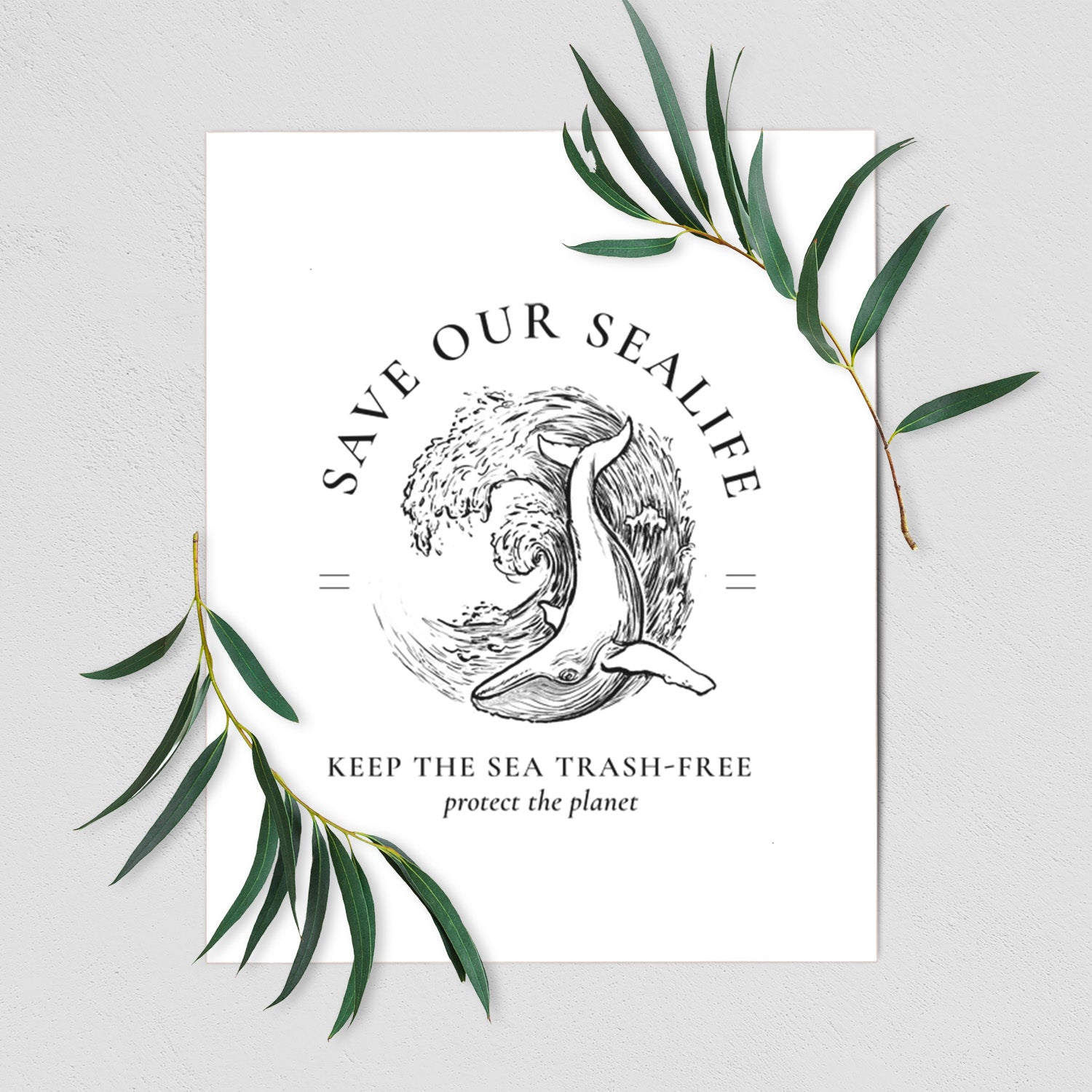 Art print of a black ink whale in an ocean wave framed by the words Save Our Sealife, Keep the Sea Trash-Free, shown with eucalyptus leaves.