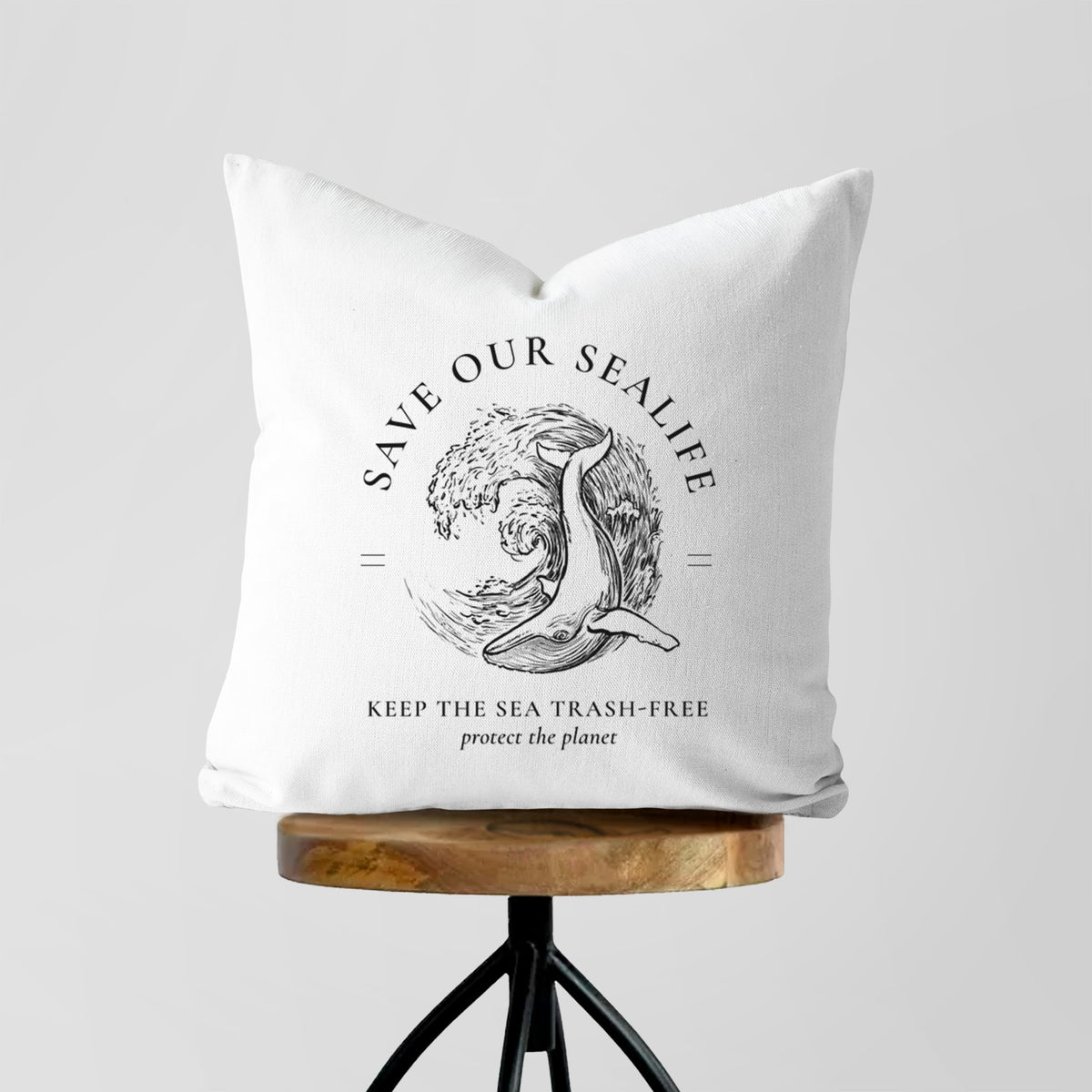 White pillow cover printed with a black ink whale in an ocean wave framed by the words Save Our Sealife, Keep the Sea Trash-Free, on a wooden stool.