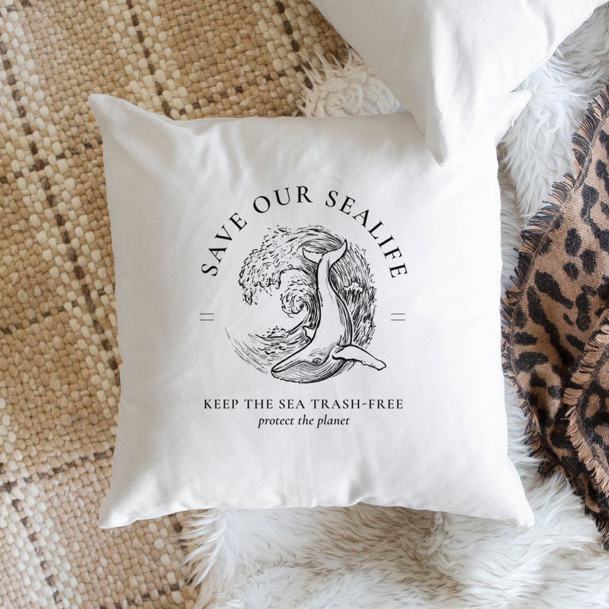 White pillow cover printed with a black ink whale in an ocean wave framed by the words Save Our Sealife, Keep the Sea Trash-Free, on a woven chair.