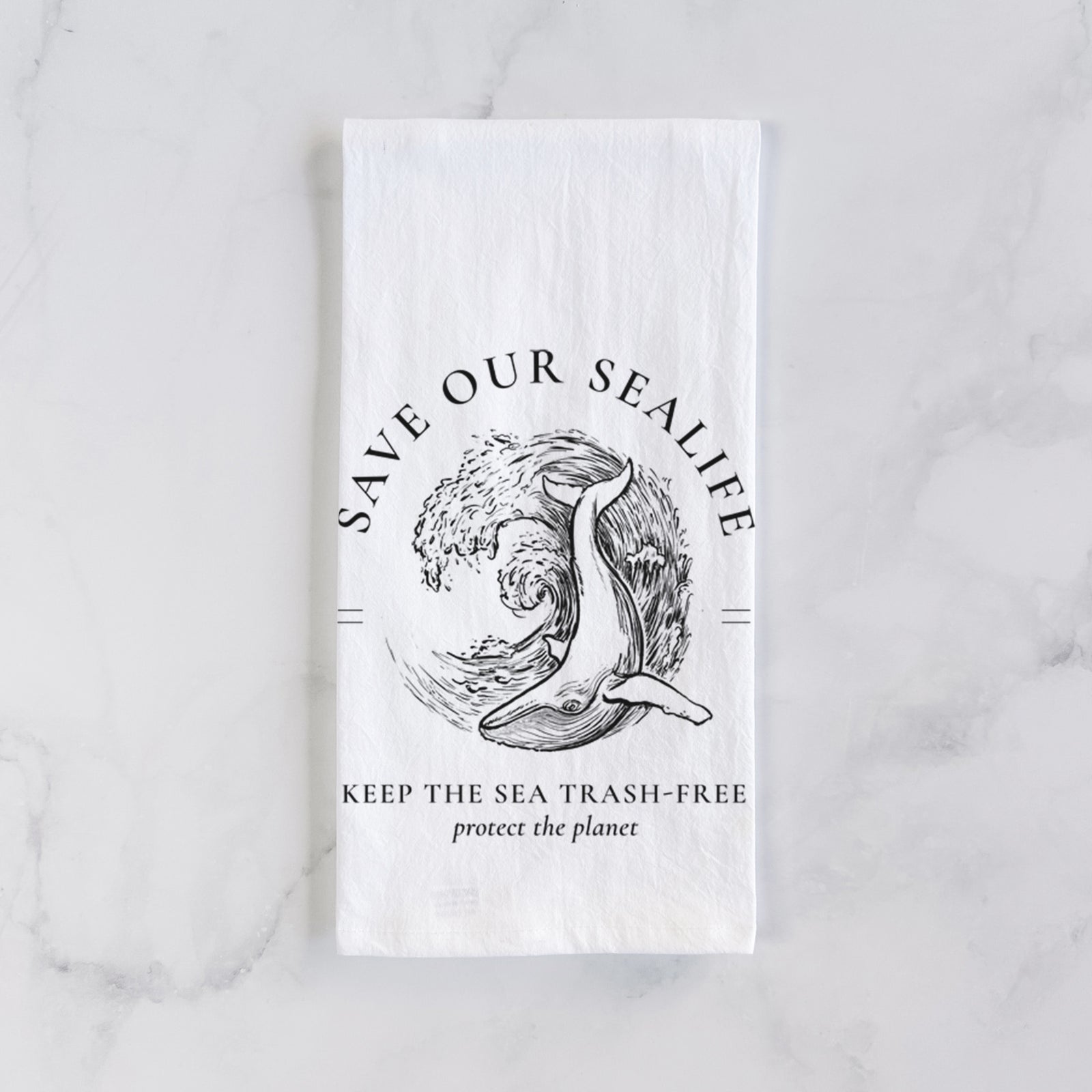 White cotton tea towel printed with a black ink whale in an ocean wave framed by the words Save Our Sealife, Keep the Sea Trash-Free, on a marble counter beside a round wooden cutting board and greenery.