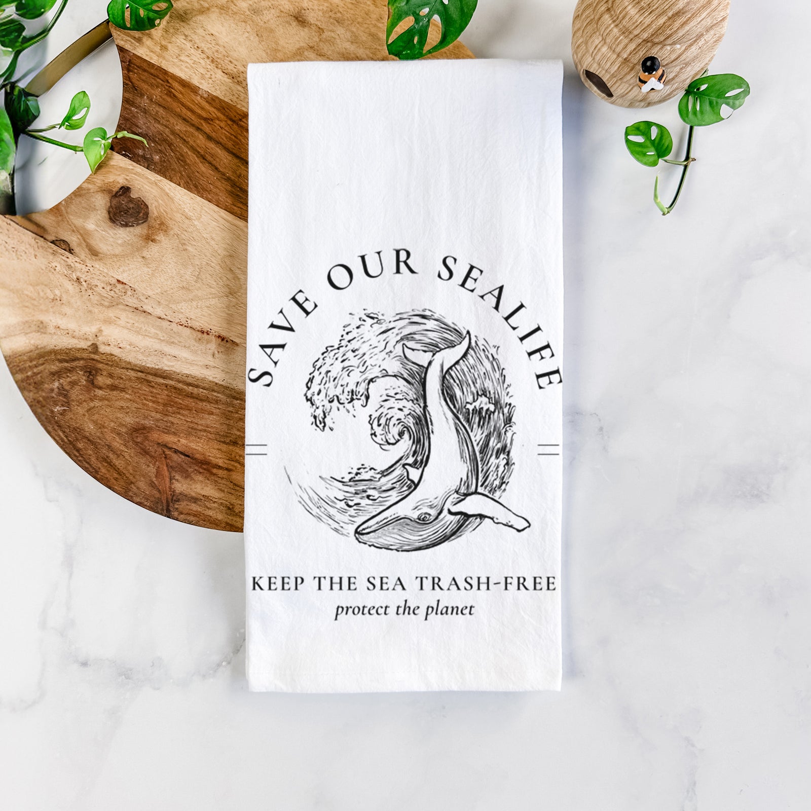 White cotton tea towel printed with a black ink whale in an ocean wave framed by the words Save Our Sealife, Keep the Sea Trash-Free, on a marble counter beside a round wooden cutting board and greenery.