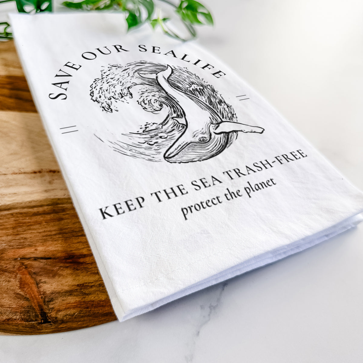 Close-up of a folded white tea towel on a wooden board, showing a black ink whale in an ocean wave framed by the words Save Our Sealife, Keep the Sea Trash-Free.