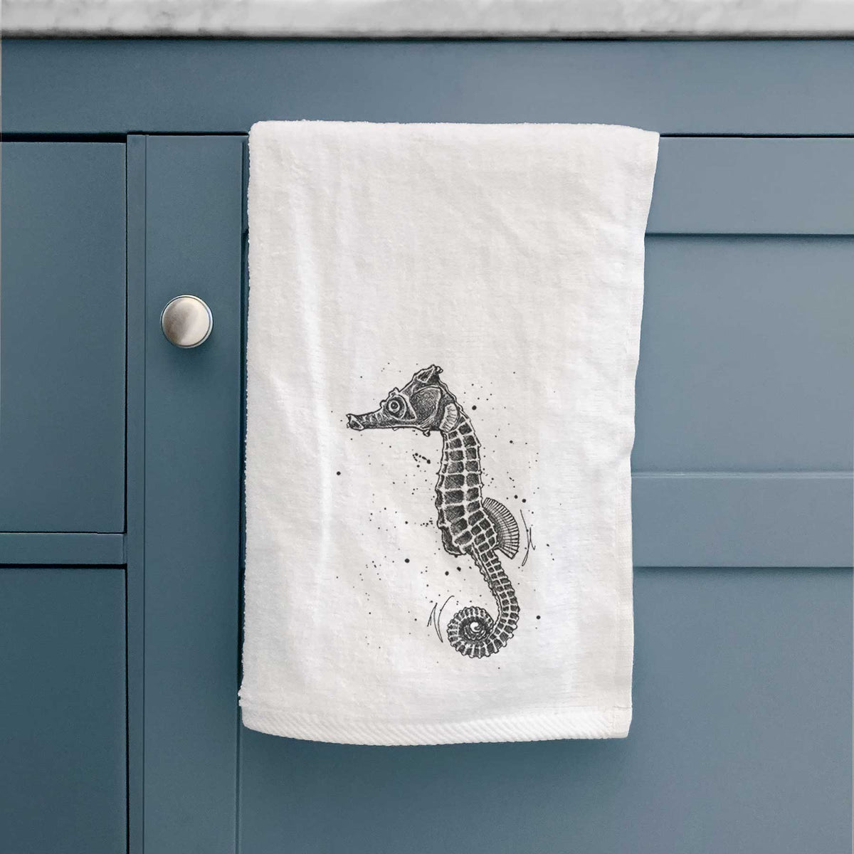 White hand towel printed with a detailed black ink illustration of a Pacific seahorse with its tail curled, hanging on a cabinet door.