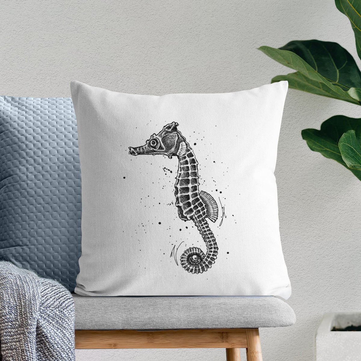 White pillow cover printed with a detailed black ink illustration of a Pacific seahorse with its tail curled, on a bench beside plants.