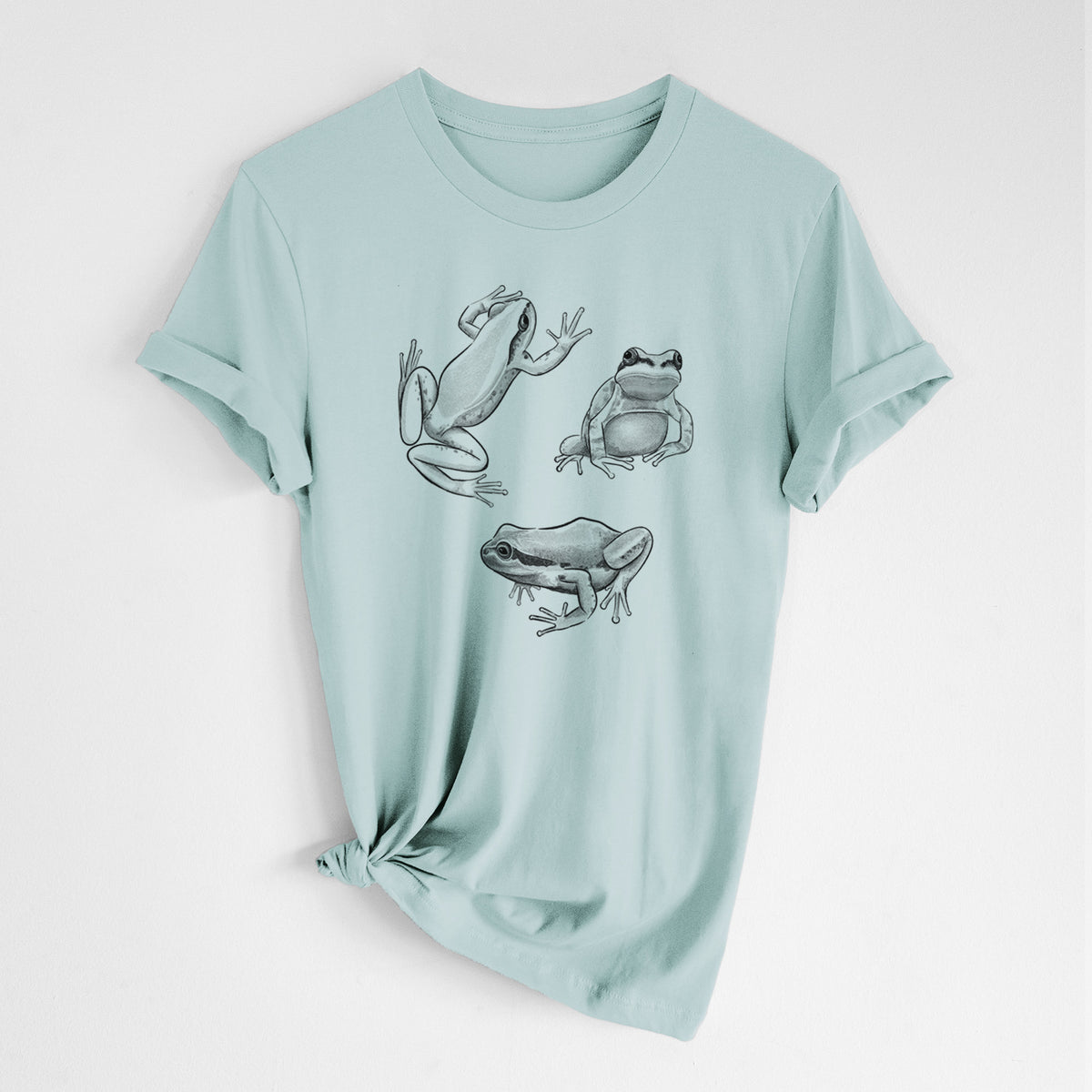 Dusty blue lightweight cotton t-shirt laid flat with the sleeves rolled, printed with a black ink illustration of three Pacific tree frogs in different poses.