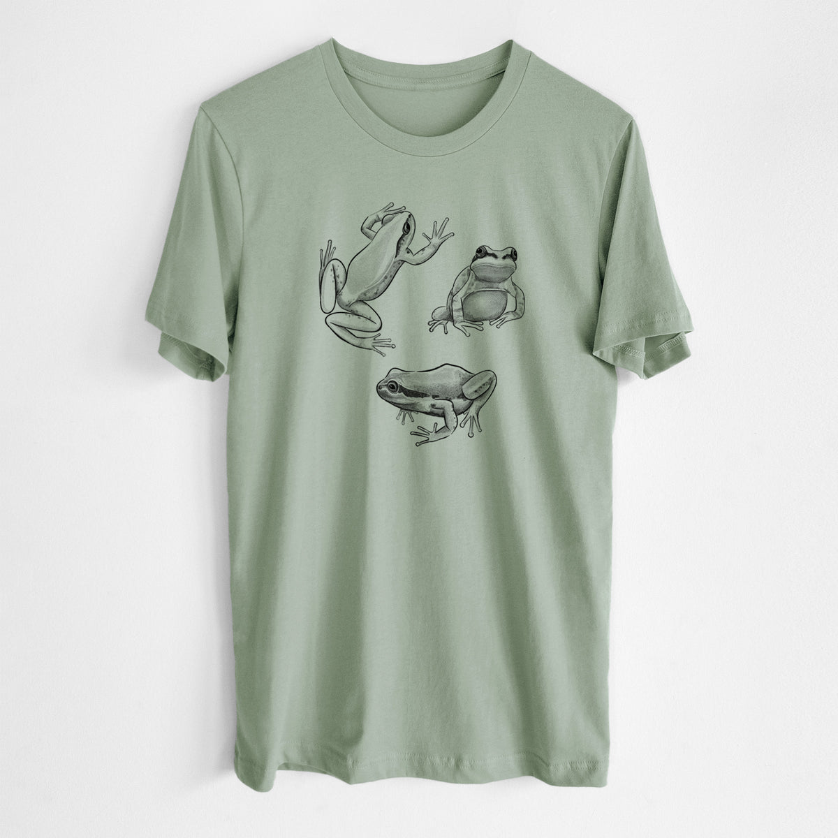 Sage green lightweight cotton t-shirt laid flat, printed with a black ink illustration of three Pacific tree frogs in different poses.