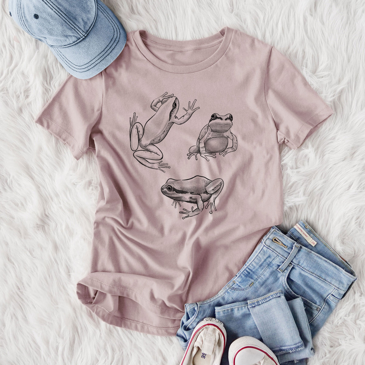 Women's relaxed fit cotton t-shirt printed with a black ink illustration of three Pacific tree frogs in different poses, styled flat on a white faux fur rug with jeans.