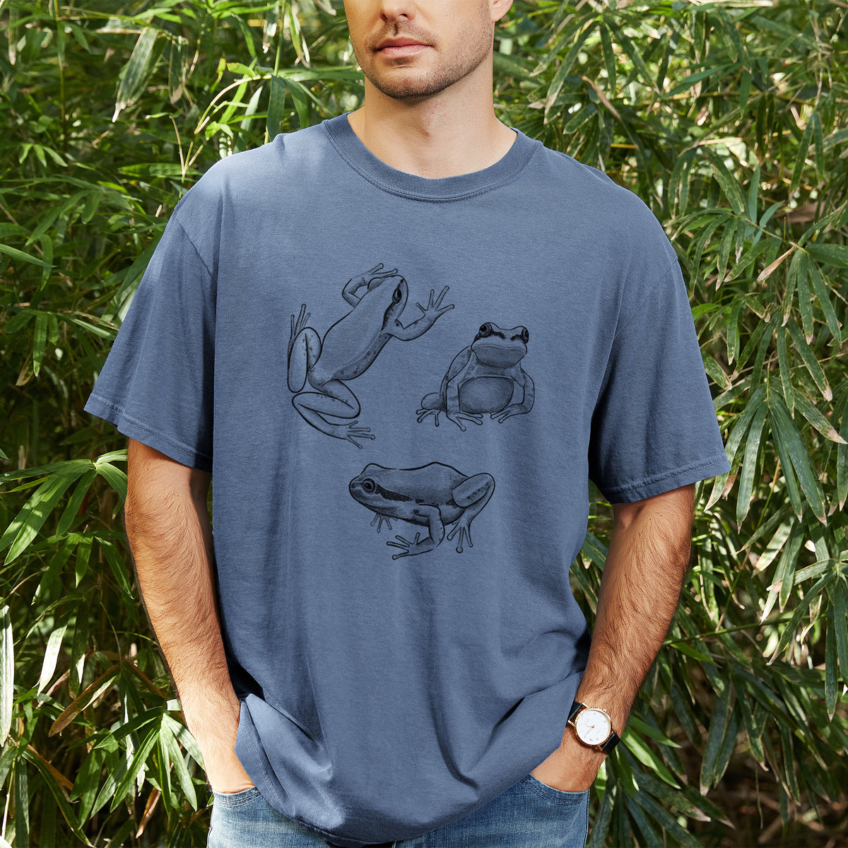 Man wearing a Comfort Colors t-shirt printed with a black ink illustration of three Pacific tree frogs in different poses, outdoors.