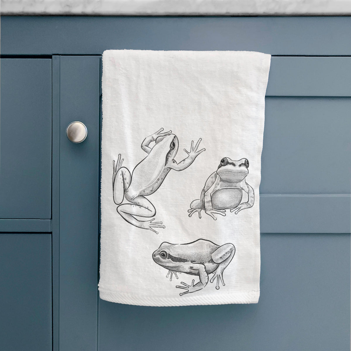 White hand towel printed with a black ink illustration of three Pacific tree frogs in different poses, hanging on a cabinet door.