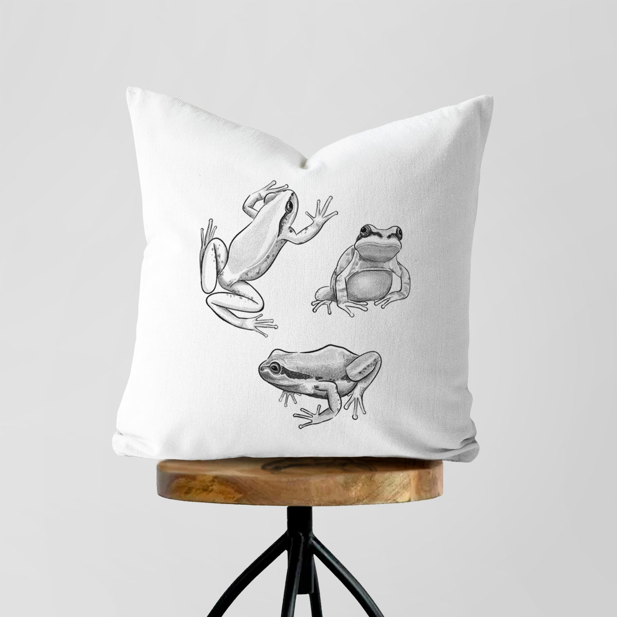 White pillow cover printed with a black ink illustration of three Pacific tree frogs in different poses, on a wooden stool.
