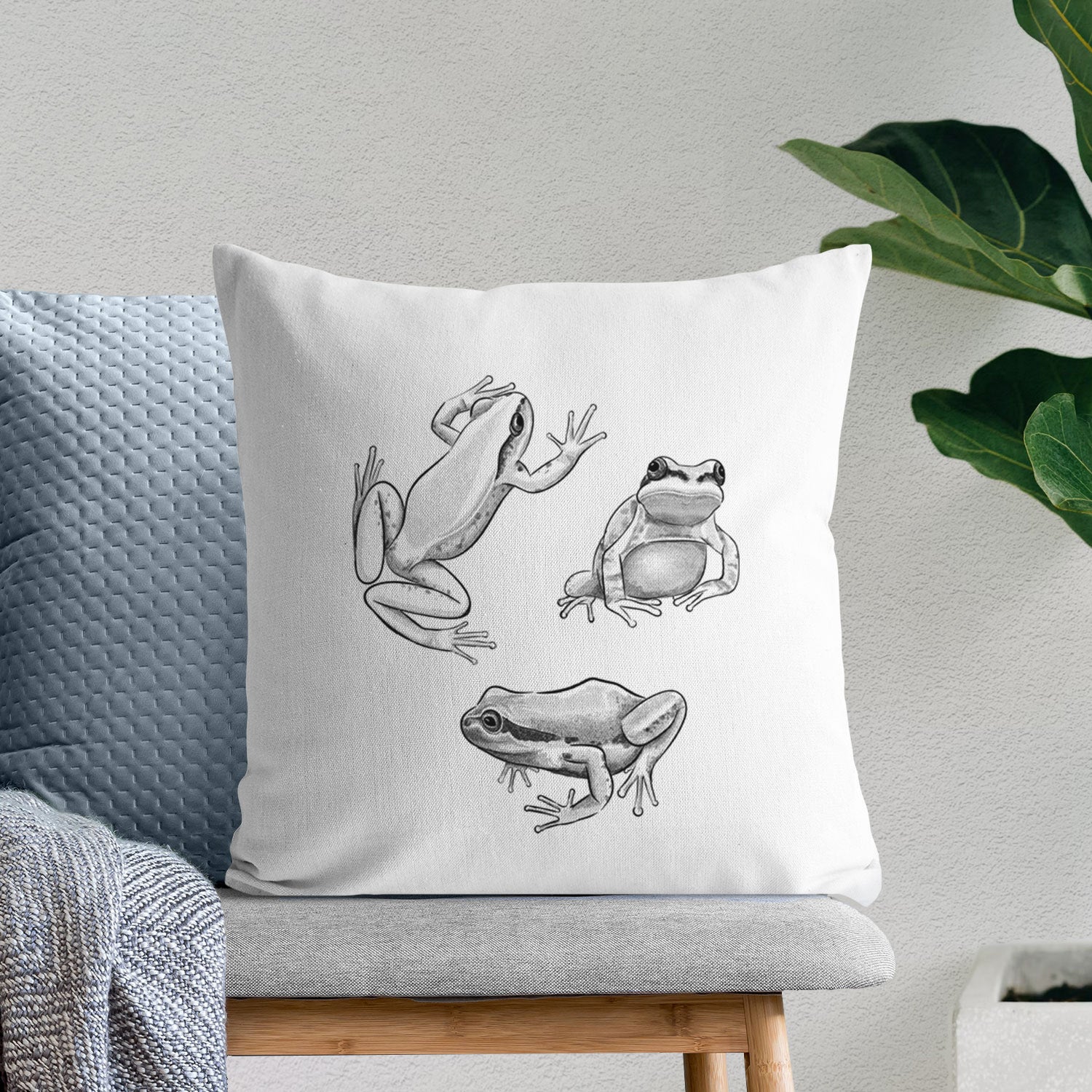 White pillow cover printed with a black ink illustration of three Pacific tree frogs in different poses, on a bench beside plants.