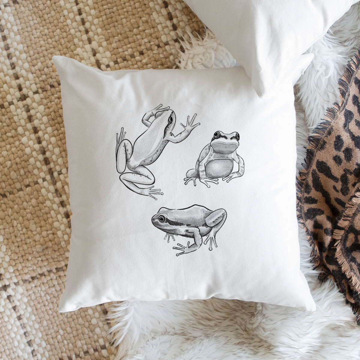 White pillow cover printed with a black ink illustration of three Pacific tree frogs in different poses, on a woven chair.