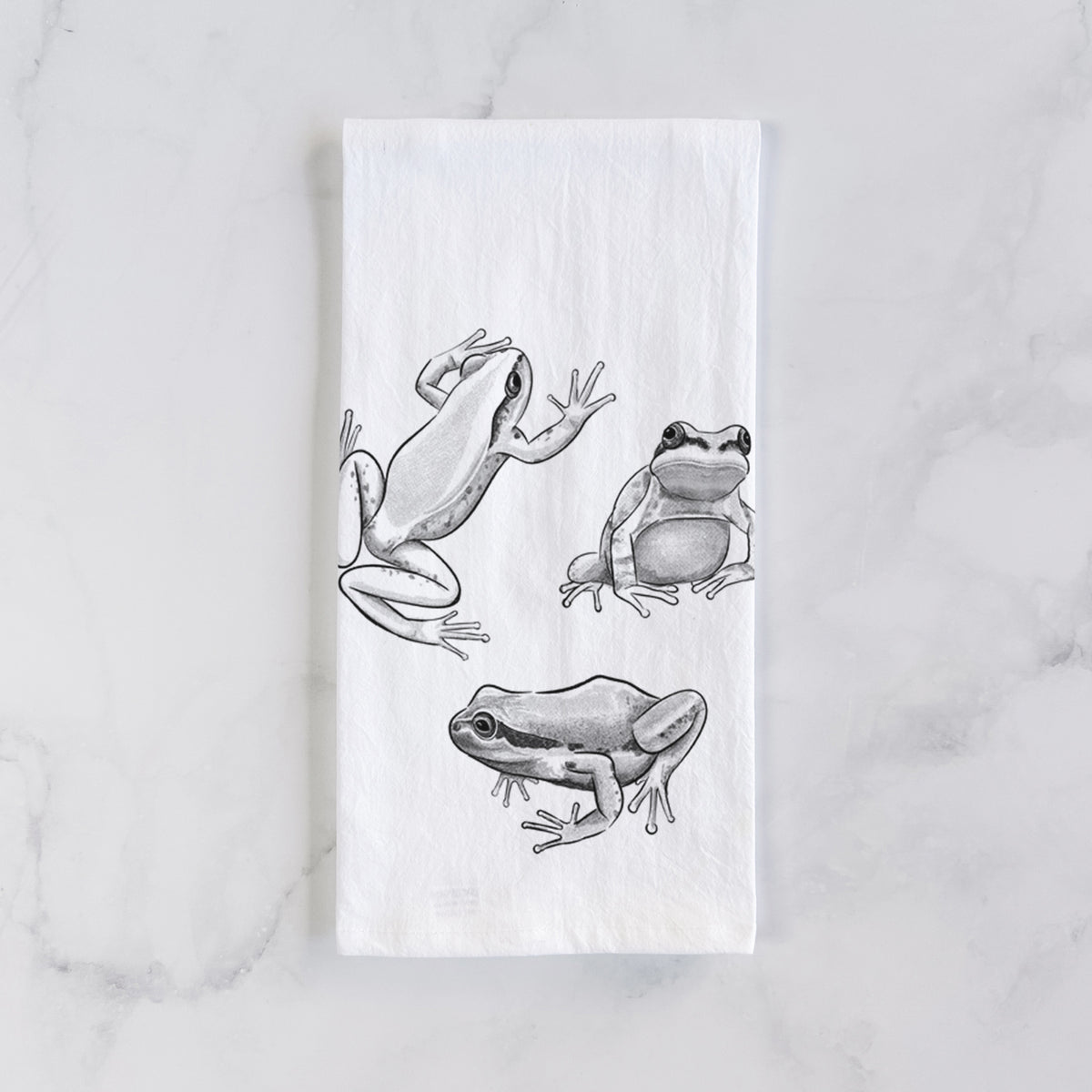 White cotton tea towel laid flat on marble, printed with a black ink illustration of three Pacific tree frogs in different poses.
