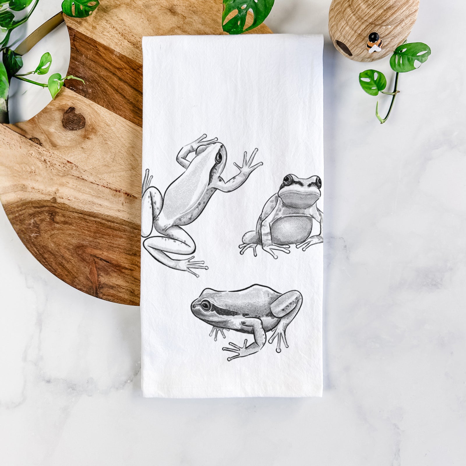 White cotton tea towel printed with a black ink illustration of three Pacific tree frogs in different poses, on a marble counter beside a round wooden cutting board and greenery.