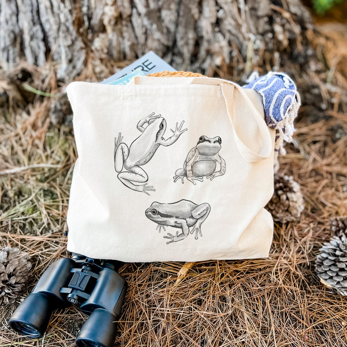 Canvas tote bag printed with a black ink illustration of three Pacific tree frogs in different poses, resting on dry grass beside a pair of binoculars.