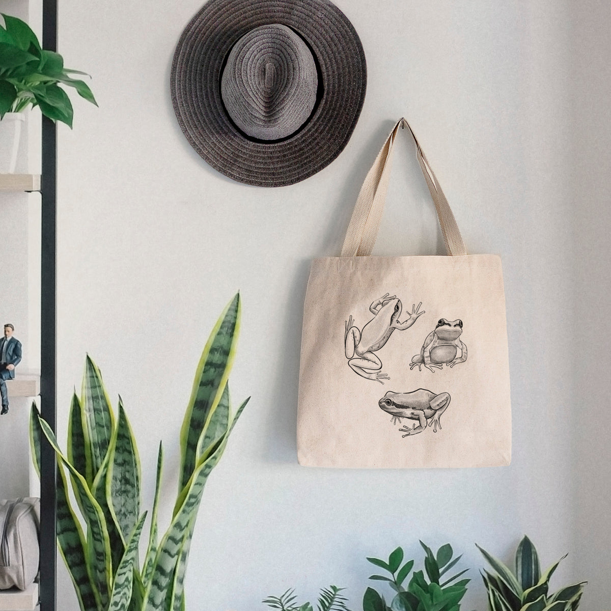 Canvas tote bag printed with a black ink illustration of three Pacific tree frogs in different poses, hanging on a wall hook below a hat, next to potted plants.