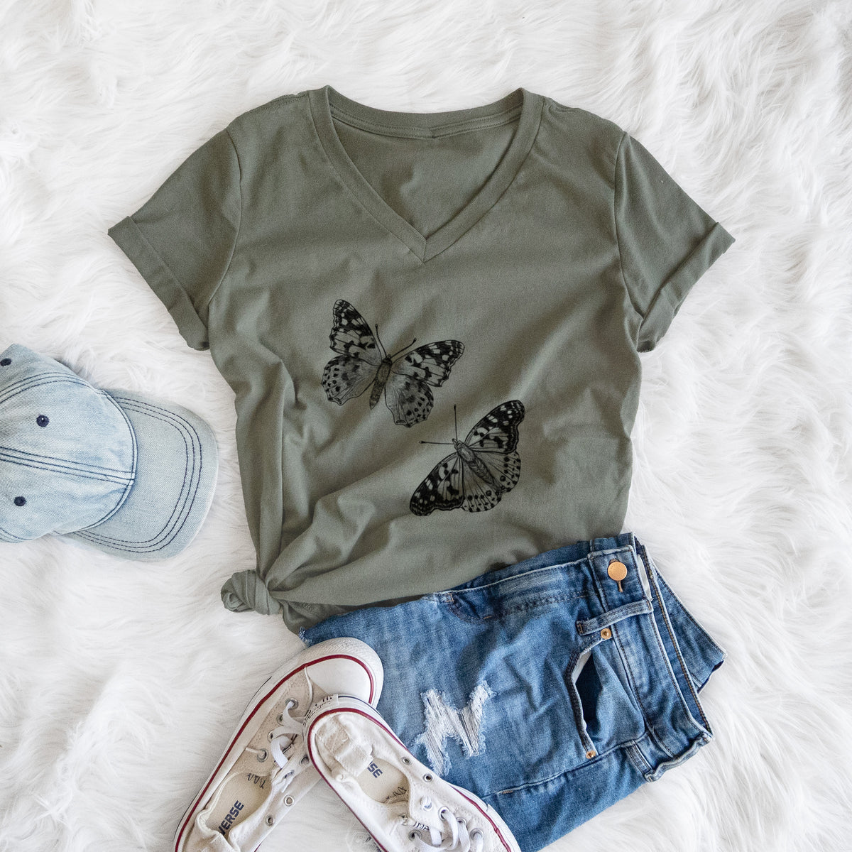 Military green women's relaxed fit cotton V-neck t-shirt printed with a detailed black ink illustration of two painted lady butterflies, styled flat on a white faux fur rug with a cap, jeans and sneakers.