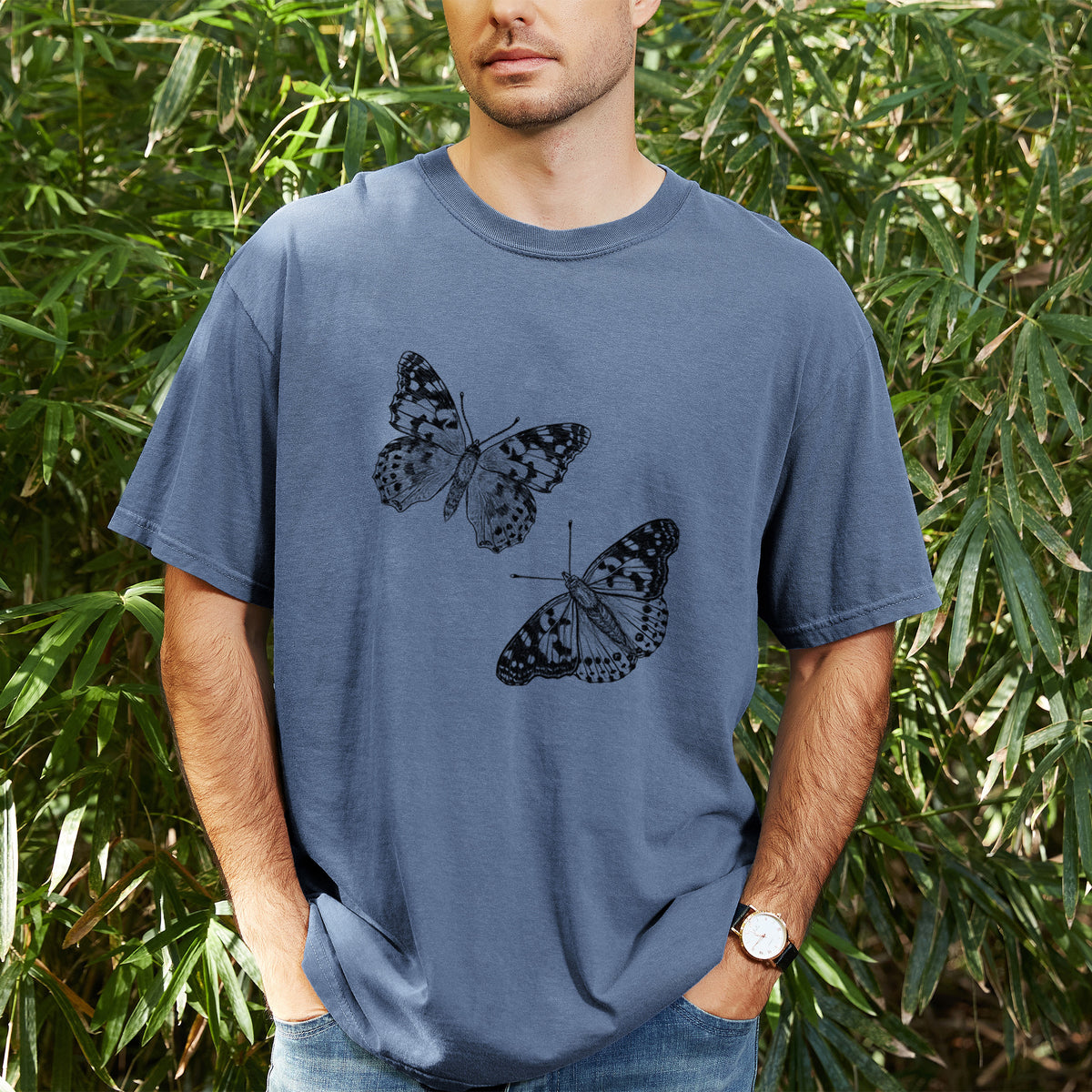 Man wearing a Comfort Colors t-shirt printed with a detailed black ink illustration of two painted lady butterflies, outdoors.