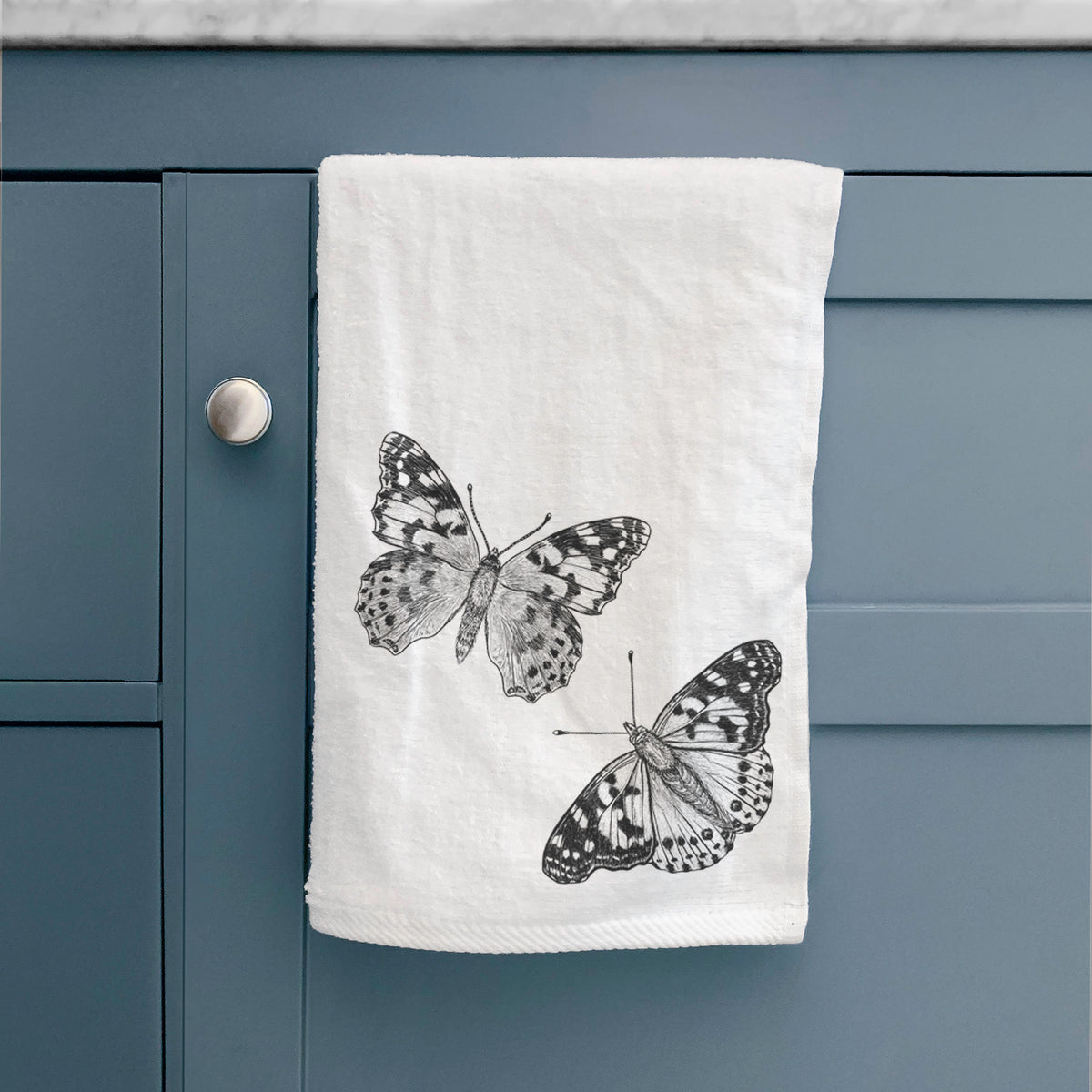 White hand towel printed with a detailed black ink illustration of two painted lady butterflies, hanging on a cabinet door.
