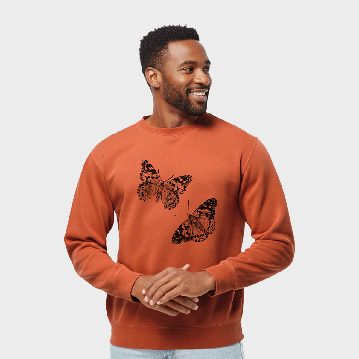 Model wearing an amber orange pigment dyed crewneck sweatshirt printed with a detailed black ink illustration of two painted lady butterflies.