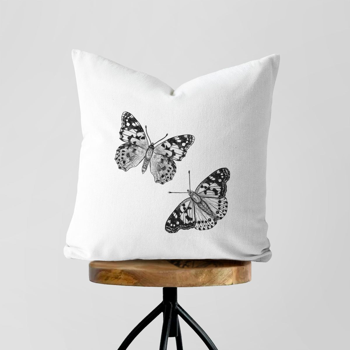 White pillow cover printed with a detailed black ink illustration of two painted lady butterflies, on a wooden stool.
