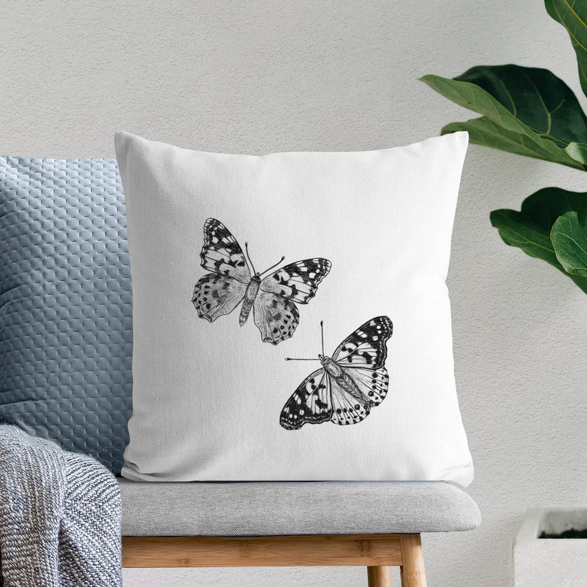 White pillow cover printed with a detailed black ink illustration of two painted lady butterflies, on a bench beside plants.