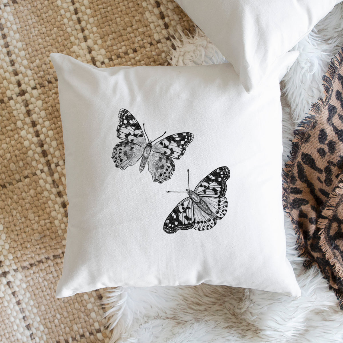 White pillow cover printed with a detailed black ink illustration of two painted lady butterflies, on a woven chair.
