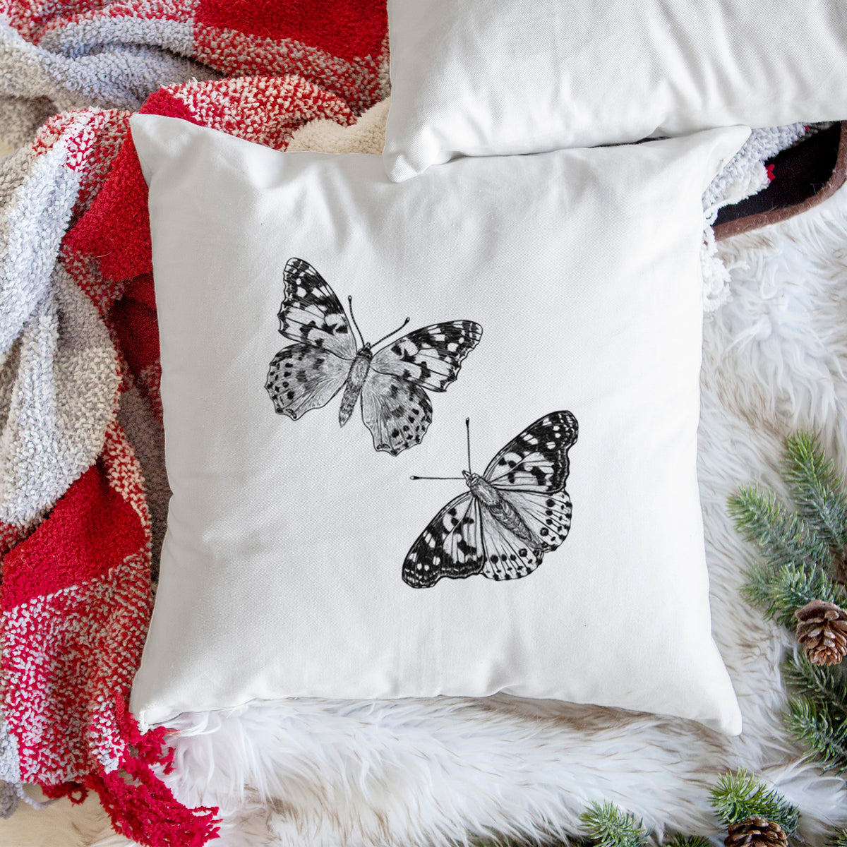White pillow cover printed with a detailed black ink illustration of two painted lady butterflies, on a woven chair.